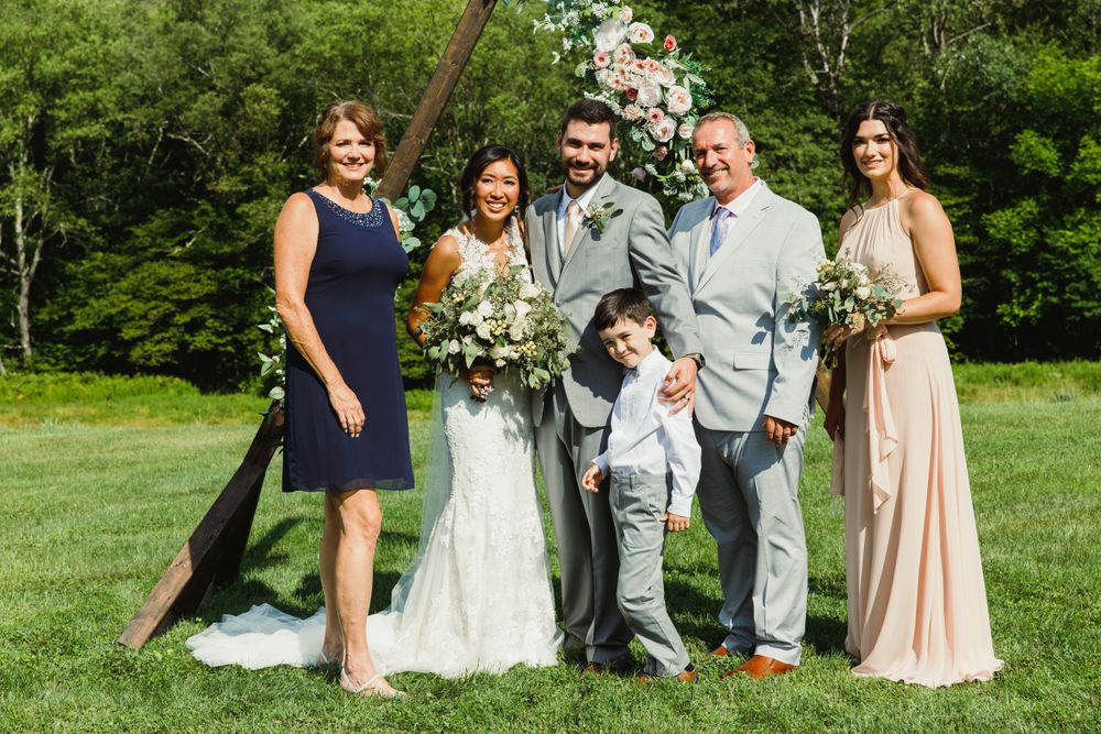 Small wedding party group photo outdoors with wooden triangle arch and forest backdrop.