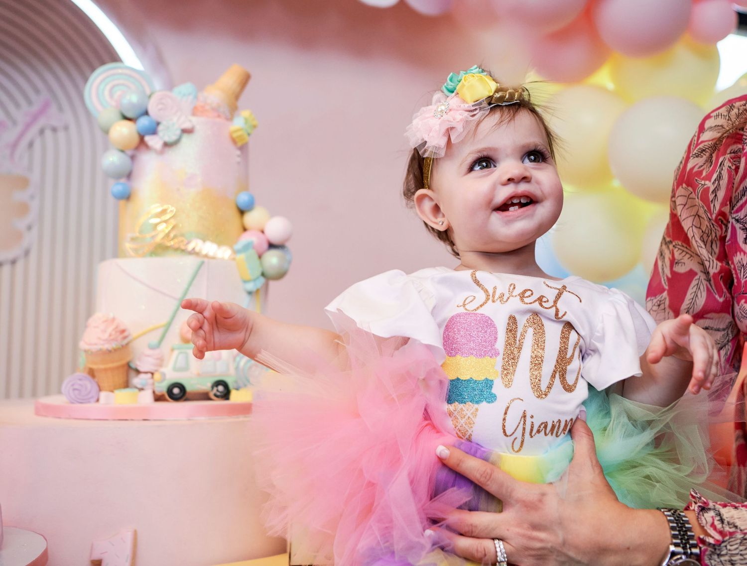 A festive first birthday party scene with a pastel cake, tulle decorations, and rainbow party details in soft pink lighting.