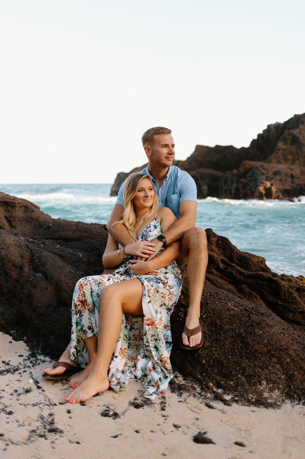 Romantic couple sits together on rocky beach overlook with crashing waves behind them.
