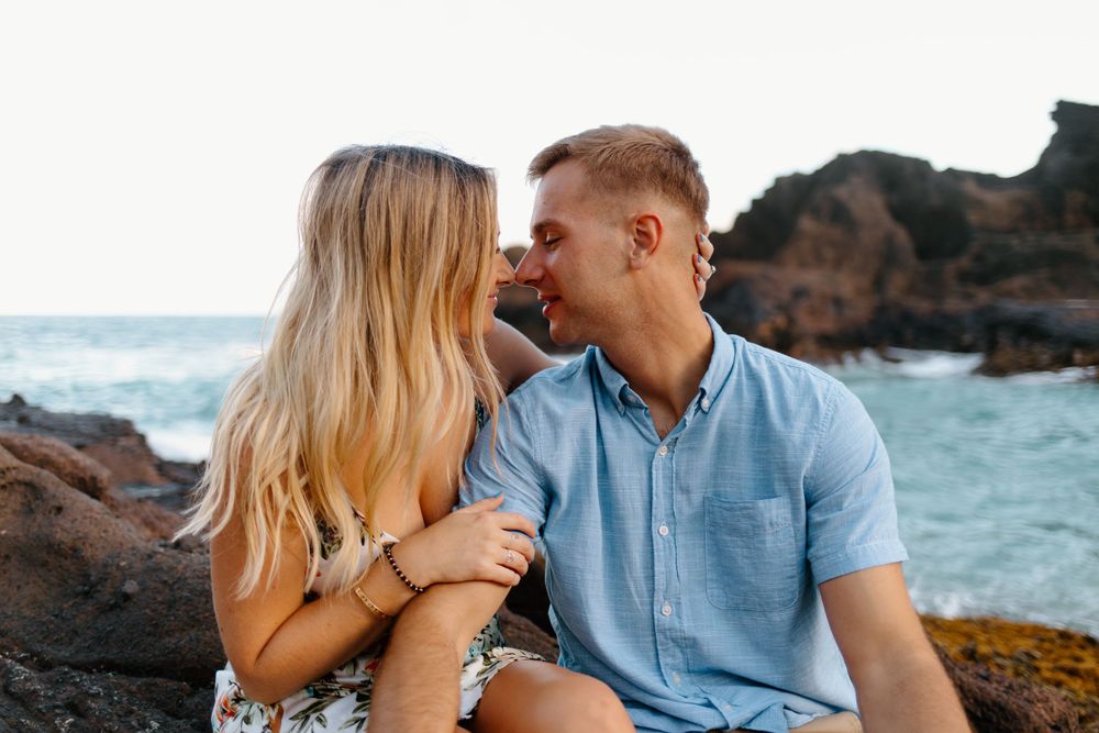 Couple sharing intimate moment on rocky coast with blue ocean waters behind them.
