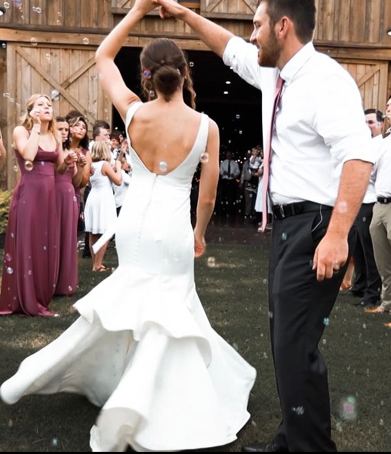 Bride in a mermaid wedding gown twirls with groom outside a rustic barn during wedding reception.