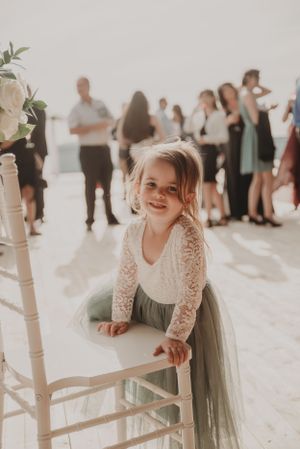 Young child in lace dress poses playfully on white chair during wedding reception.