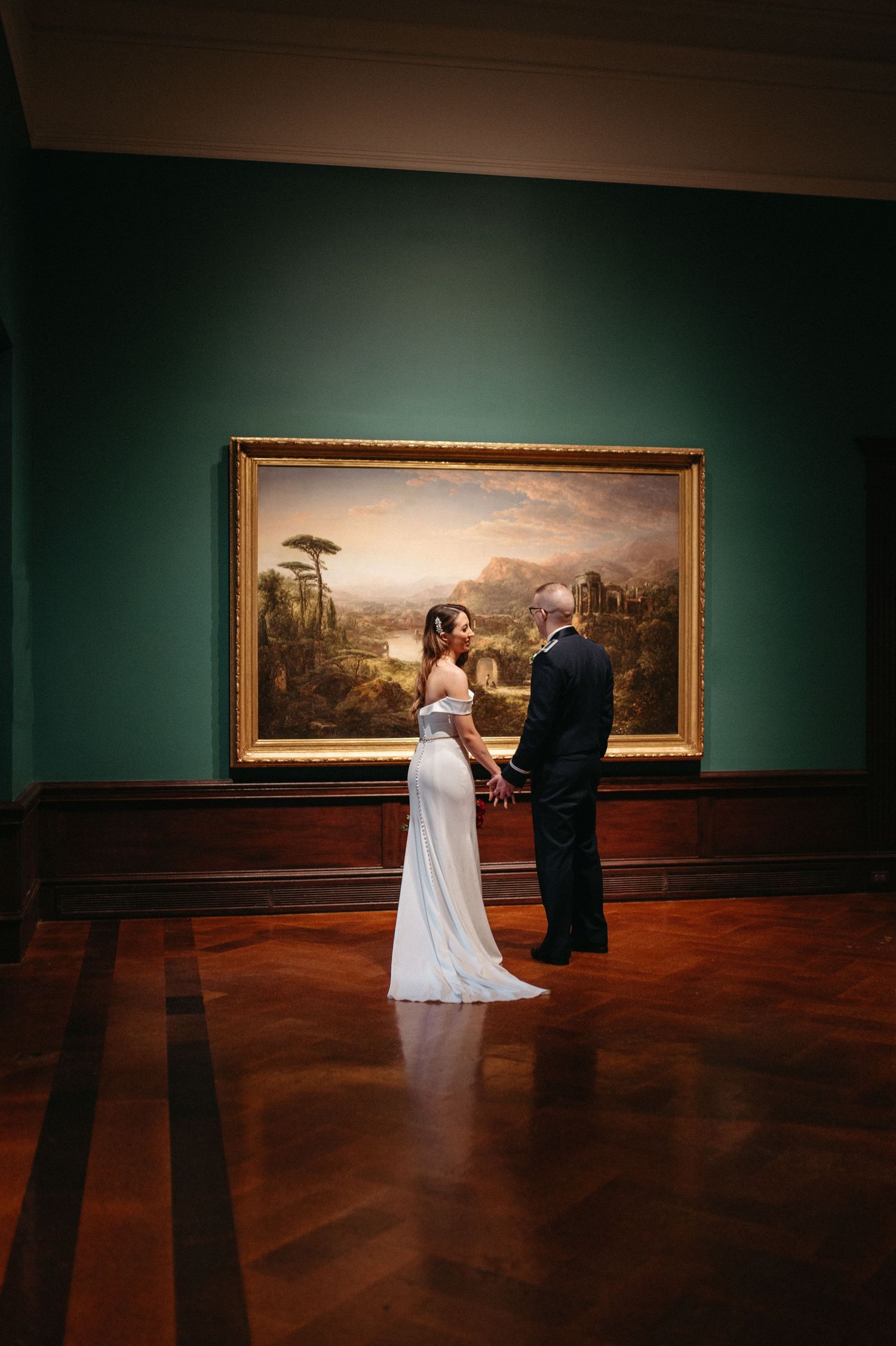 A couple in wedding attire admires a large framed painting in an elegant museum gallery.