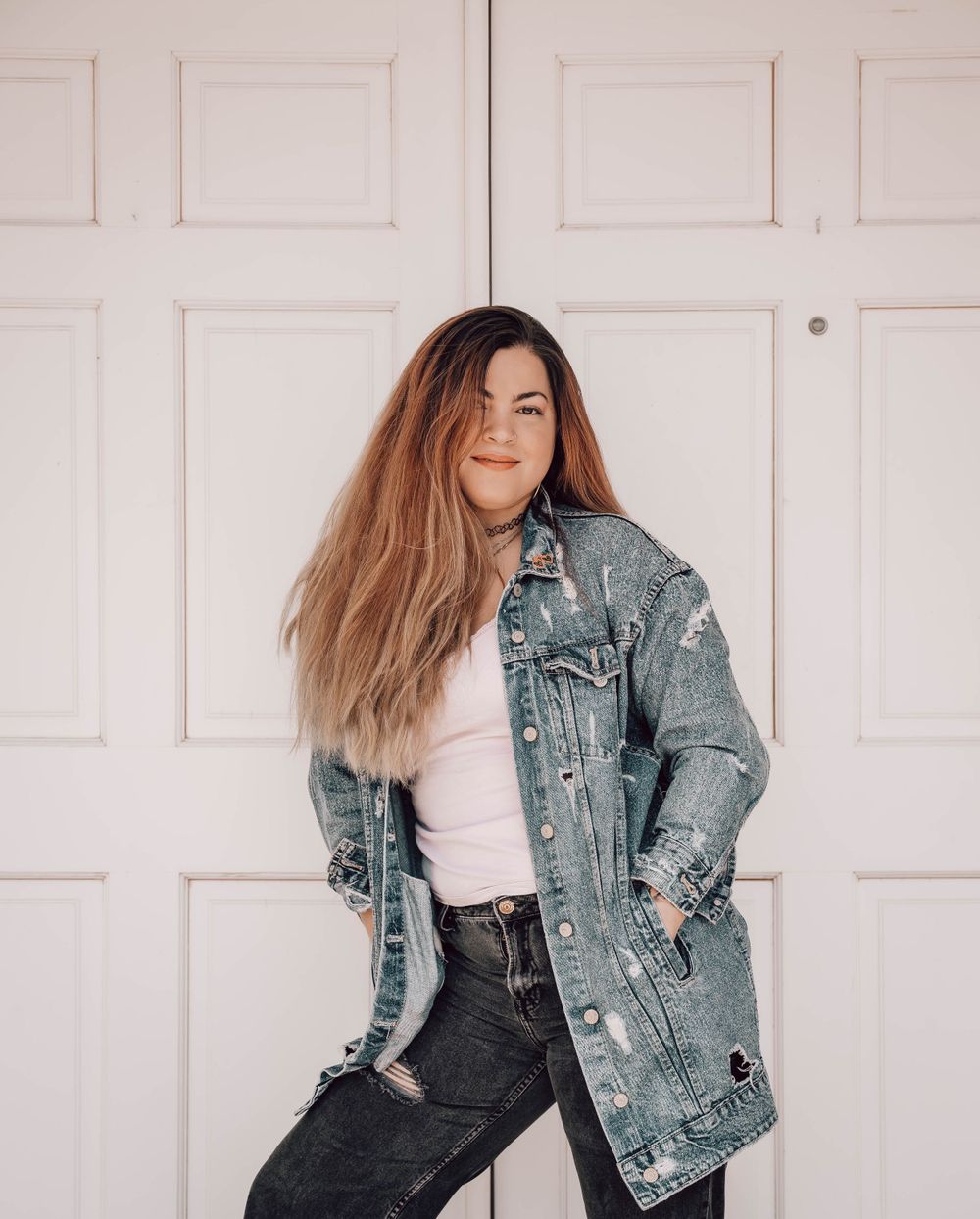 A fashion portrait against a white door featuring an oversized distressed denim jacket and casual pose.