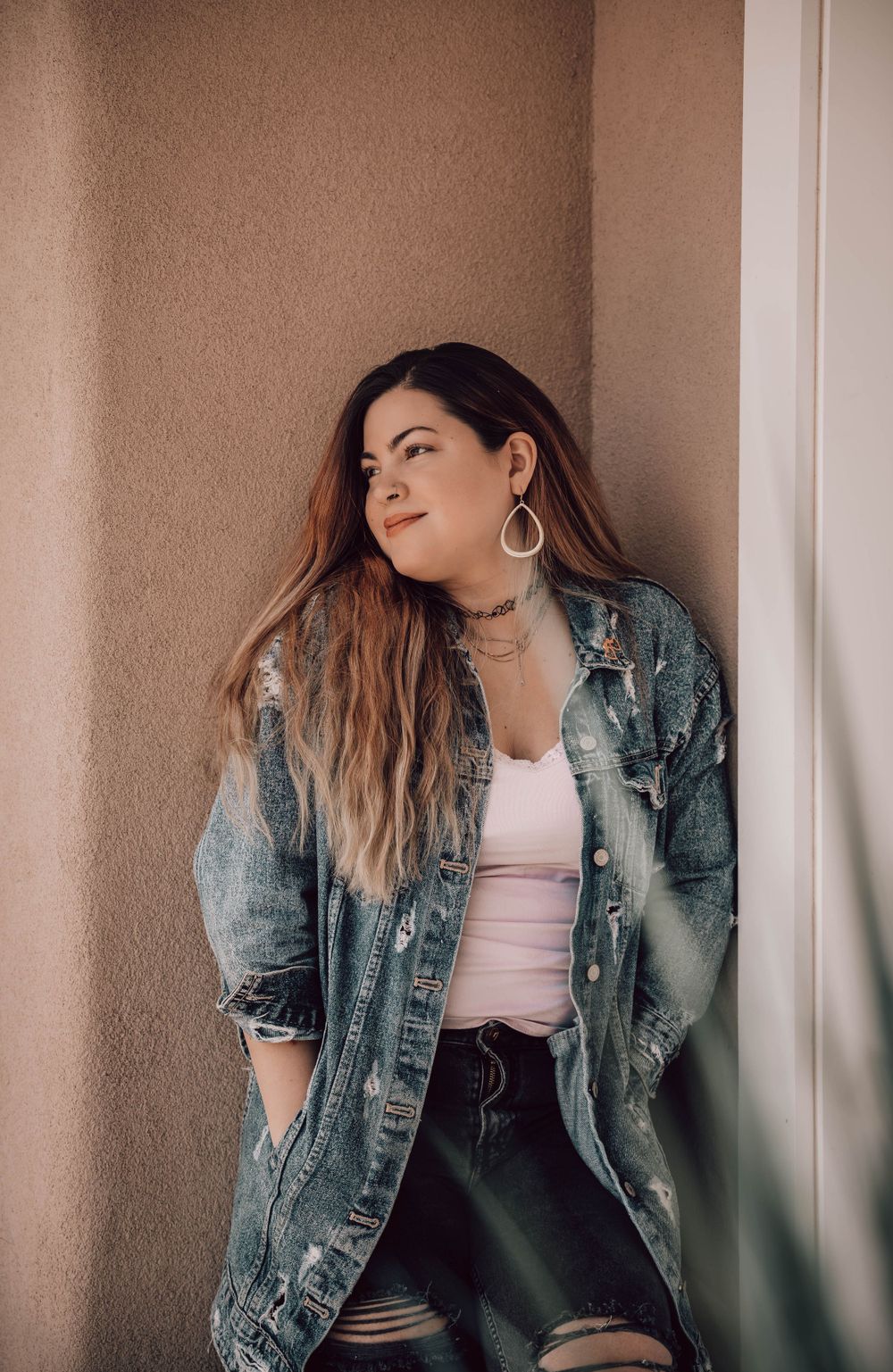 Casual fashion portrait with a distressed denim jacket and hoop earrings against a textured wall.