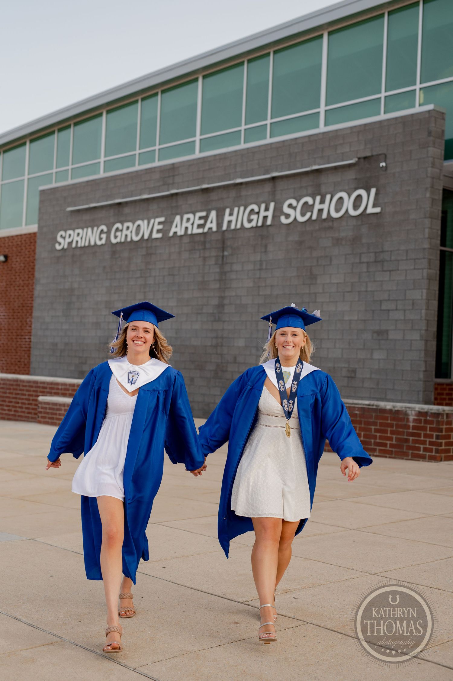 Two graduates in blue caps and gowns holding hands while walking outside their high school.