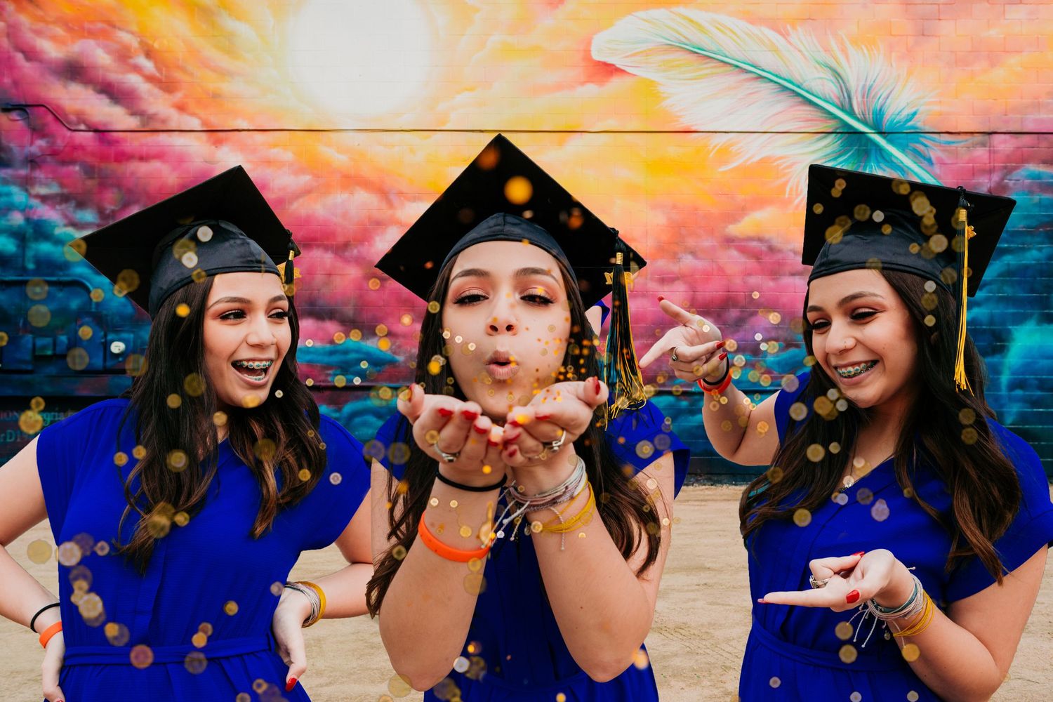 Three graduates in blue caps and gowns pose playfully against a vibrant sunset-colored wall with glitter in the air.