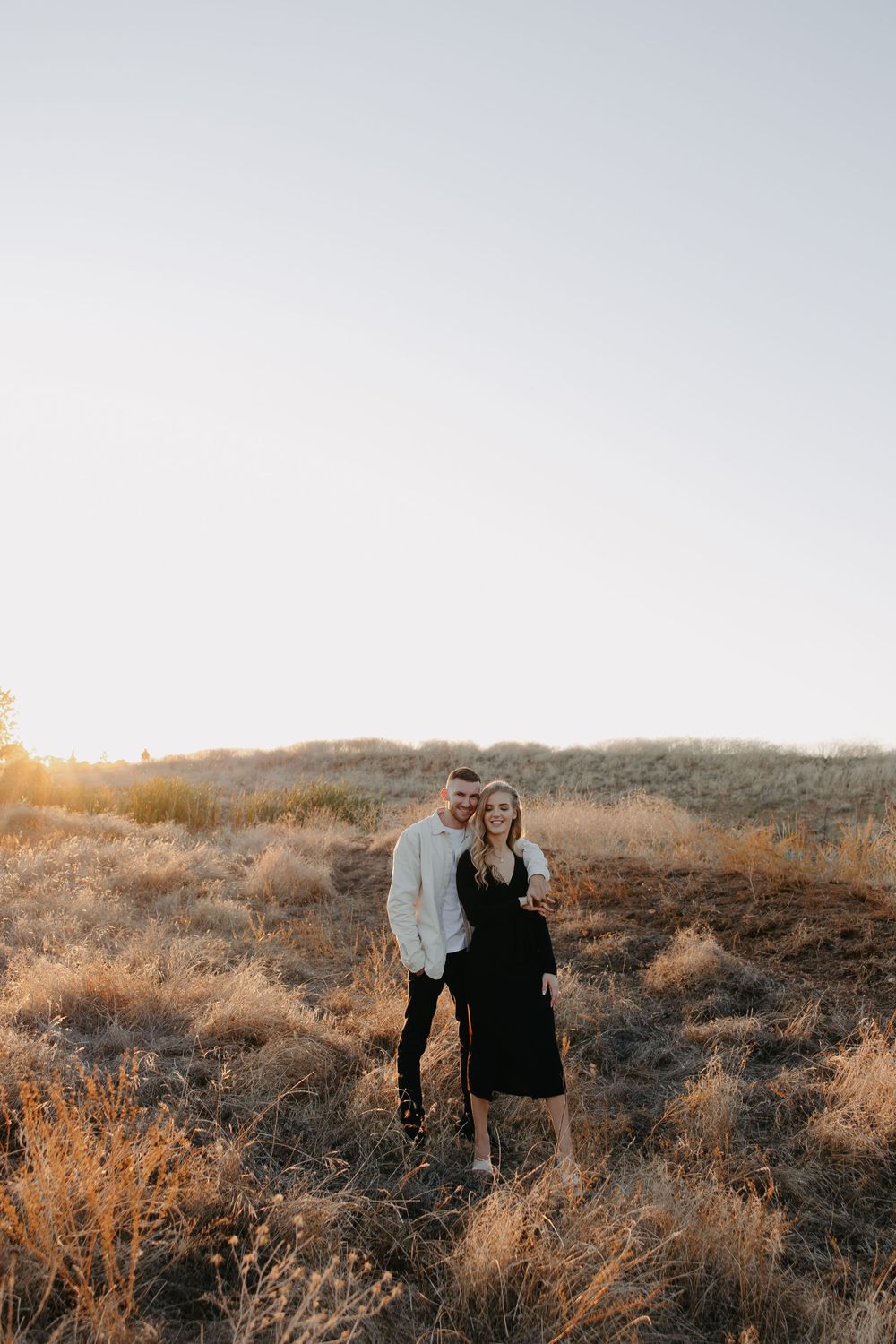 A couple poses together on a hillside during golden hour in autumn amongst dried grass and brush.