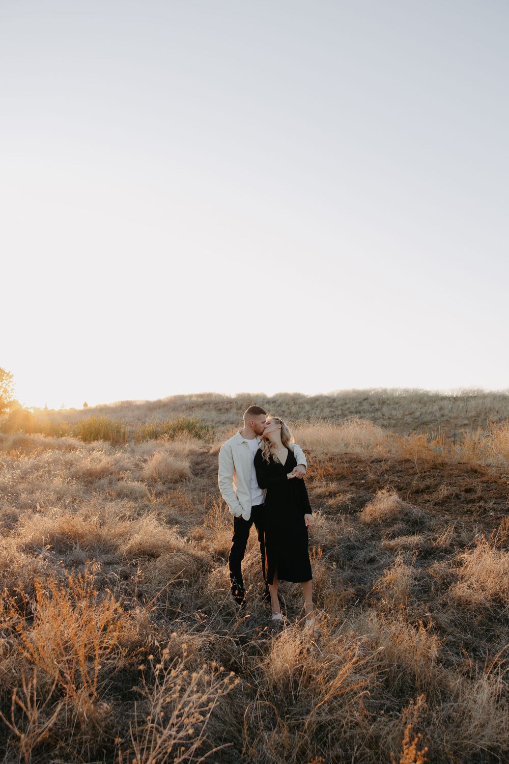 A romantic couple stands close together on a desert hillside at sunset.