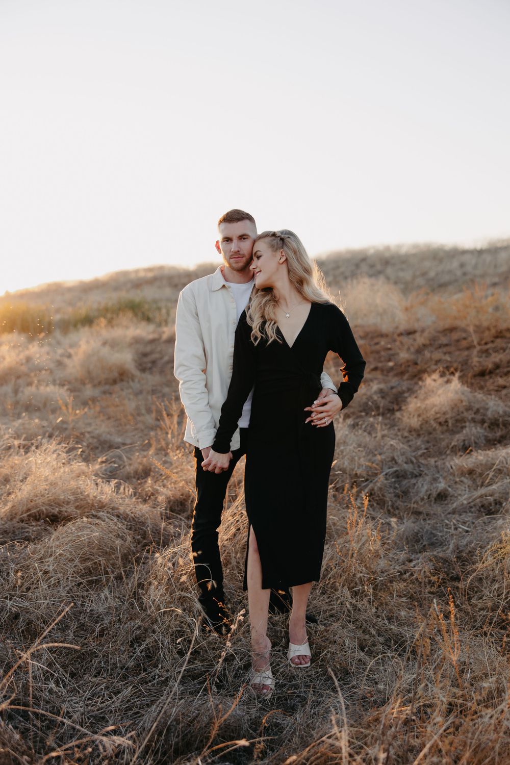 A couple in formal black and white attire poses together on a rustic hillside.