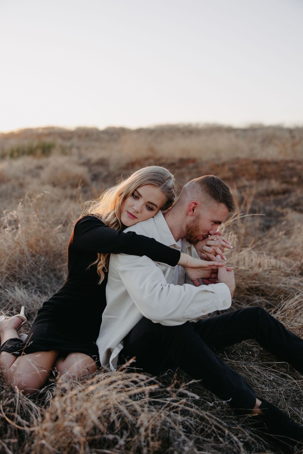 A couple embraces while sitting among dried grass on a hillside during sunset.