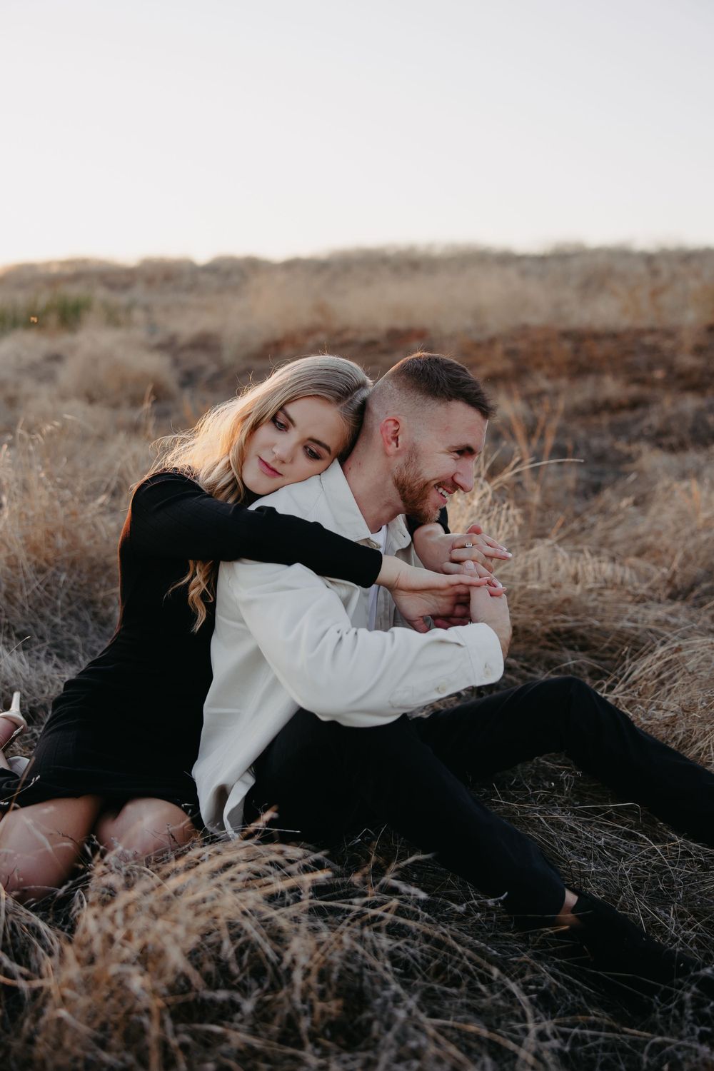 A couple shares laughter while sitting together on a golden hillside at sunset.