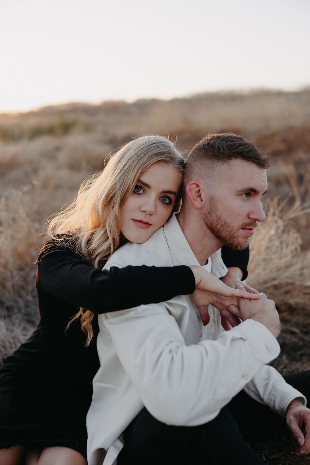 A romantic couple embraces while sitting in dry grass during golden hour sunset.