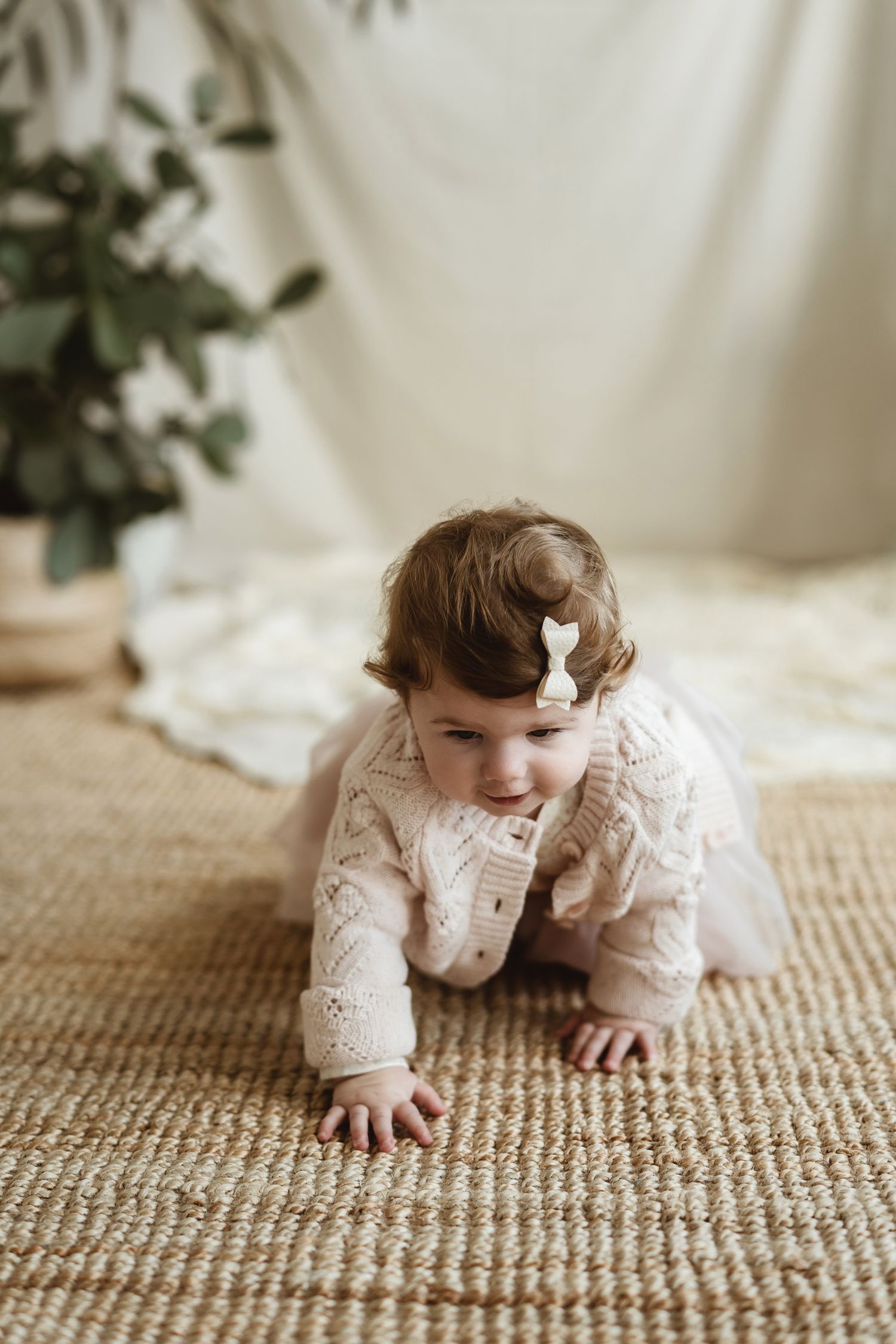 Small child in delicate white lace dress crawling on textured beige rug with white backdrop and greenery.