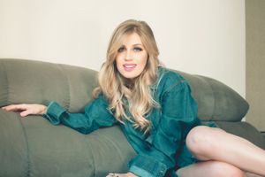 Fashion portrait on a grey couch wearing a teal button-up dress with blonde wavy hair.