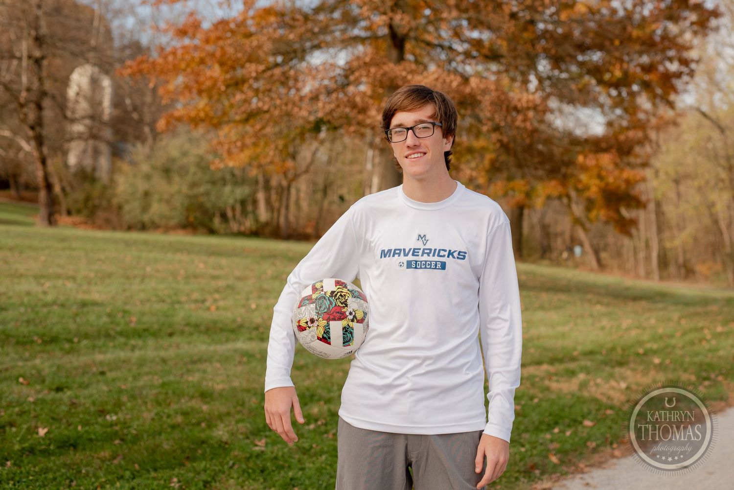 Student athlete in white long sleeve shirt poses in park with autumn trees.