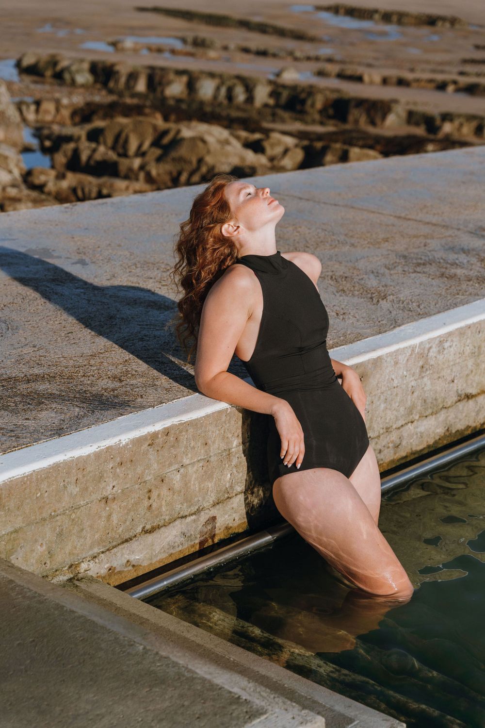 Woman in black swimsuit relaxing at poolside with water in background.