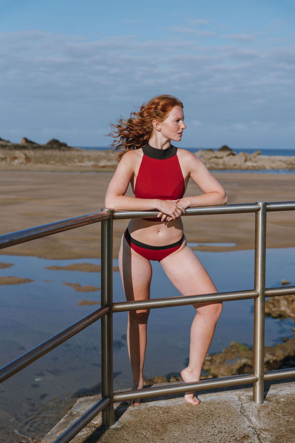 Athletic figure in red swimwear poses along coastal railing with ocean and rocks in background.