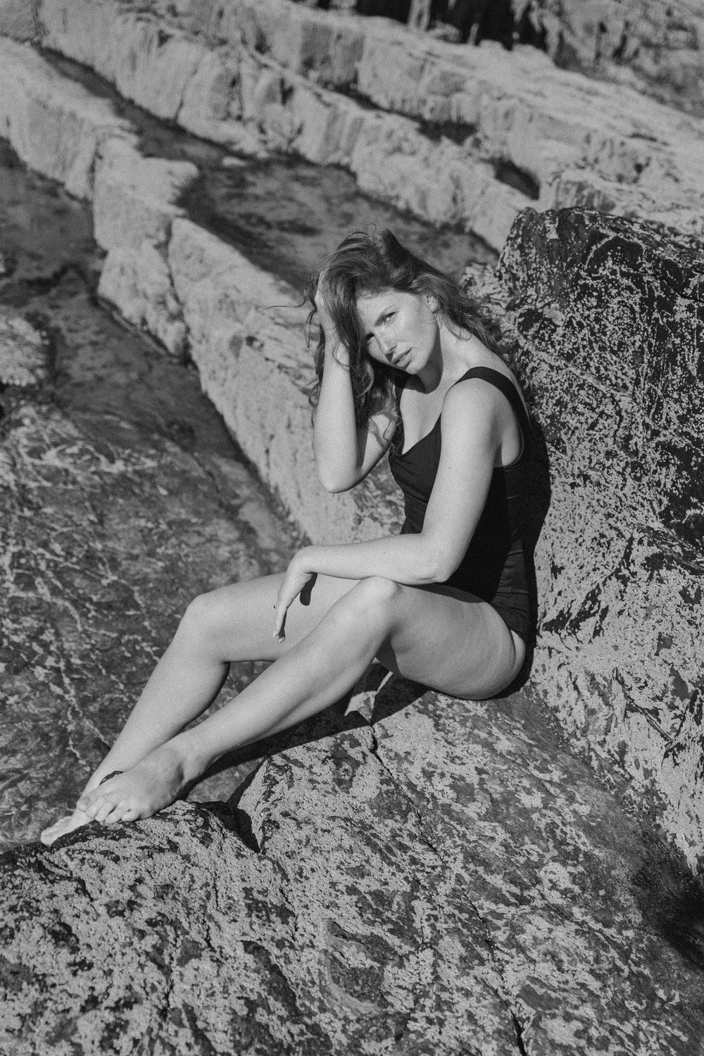 Black and white photo of figure in swimsuit posing on rocky coastal outcrop.