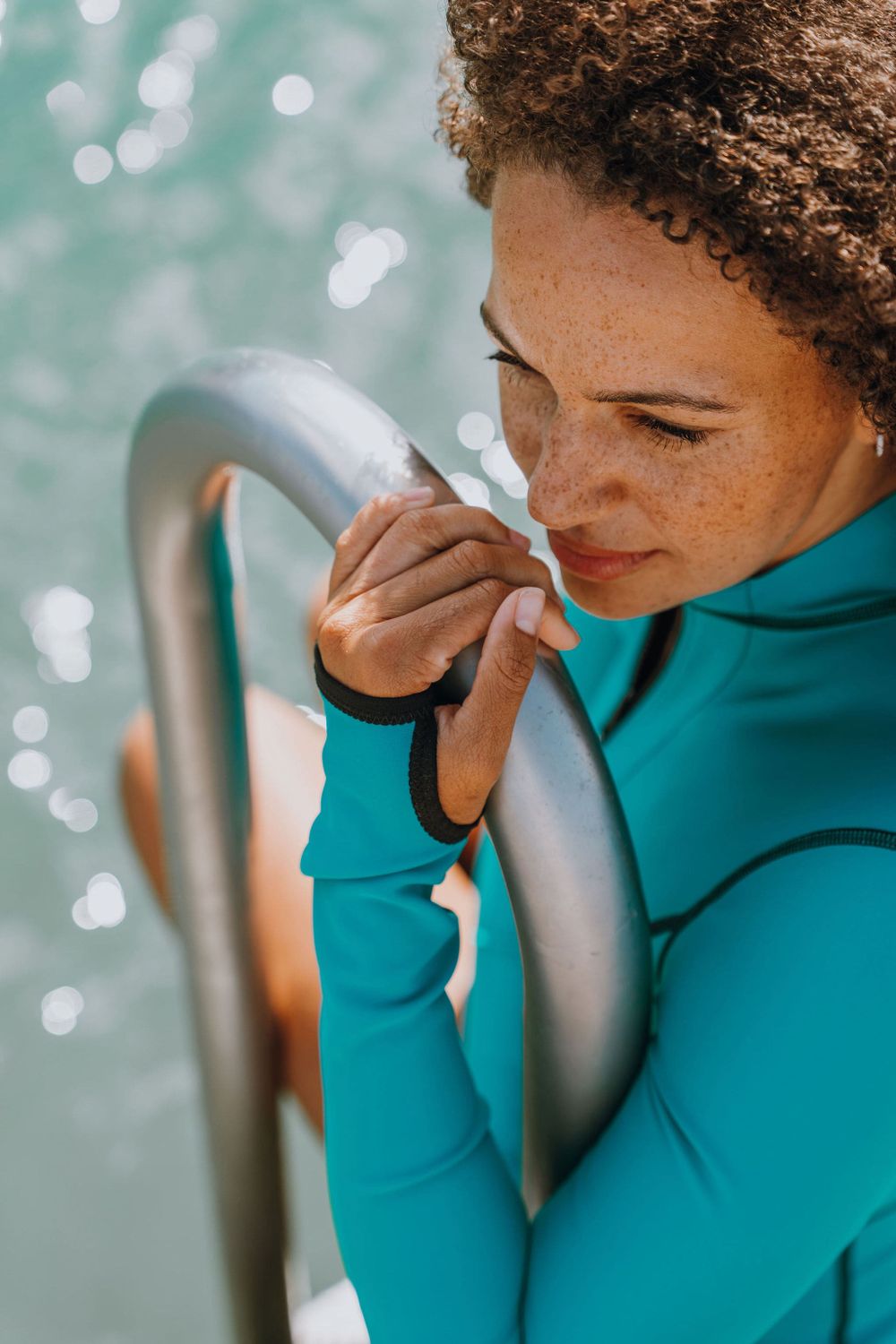 Close-up of person in teal wetsuit holding pool railing with sparkling water in background.