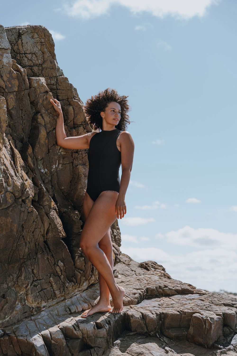 Model in black swimsuit poses confidently against dramatic rocky cliff face.