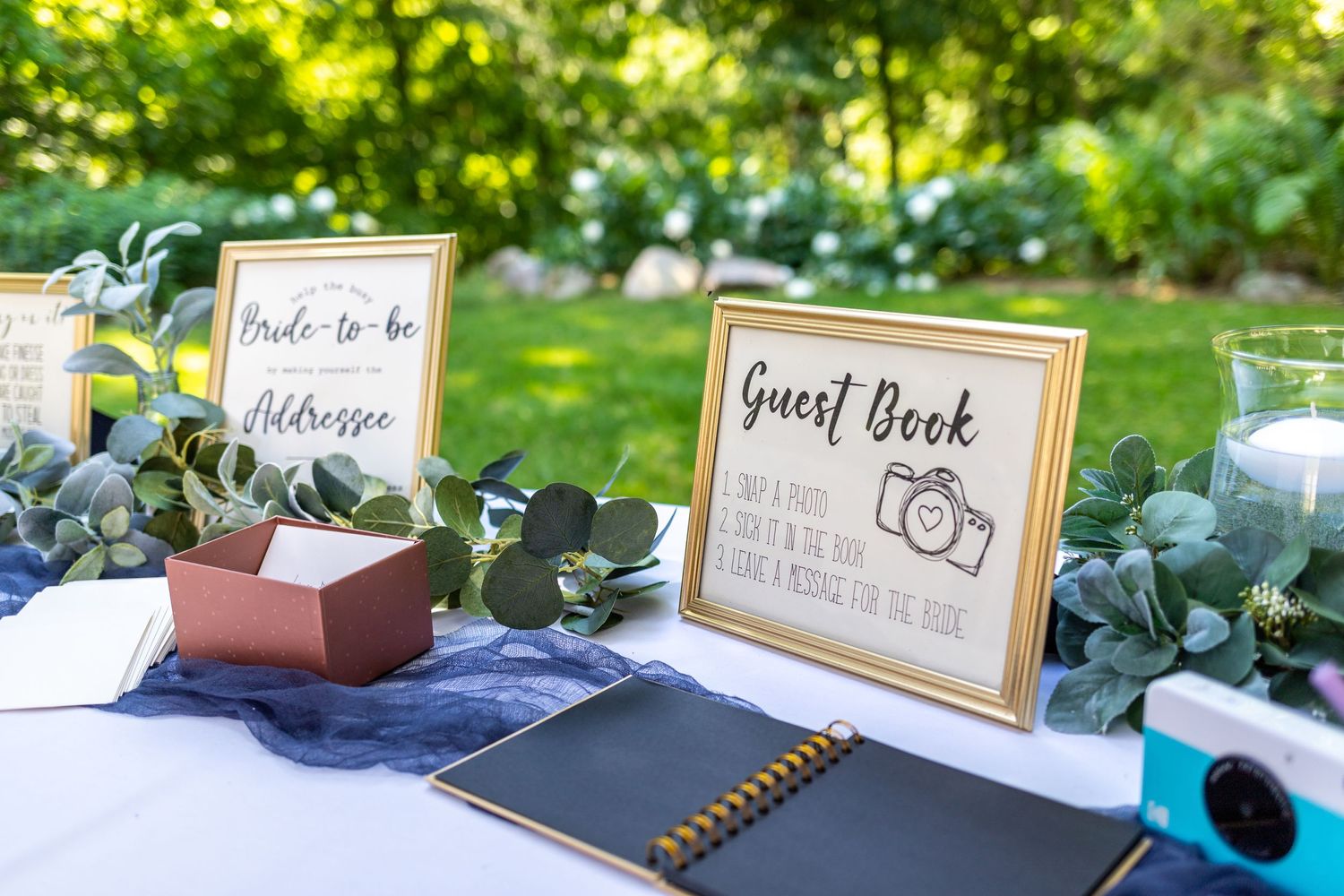 Outdoor wedding guest book table decorated with eucalyptus greenery and gold framed signs.