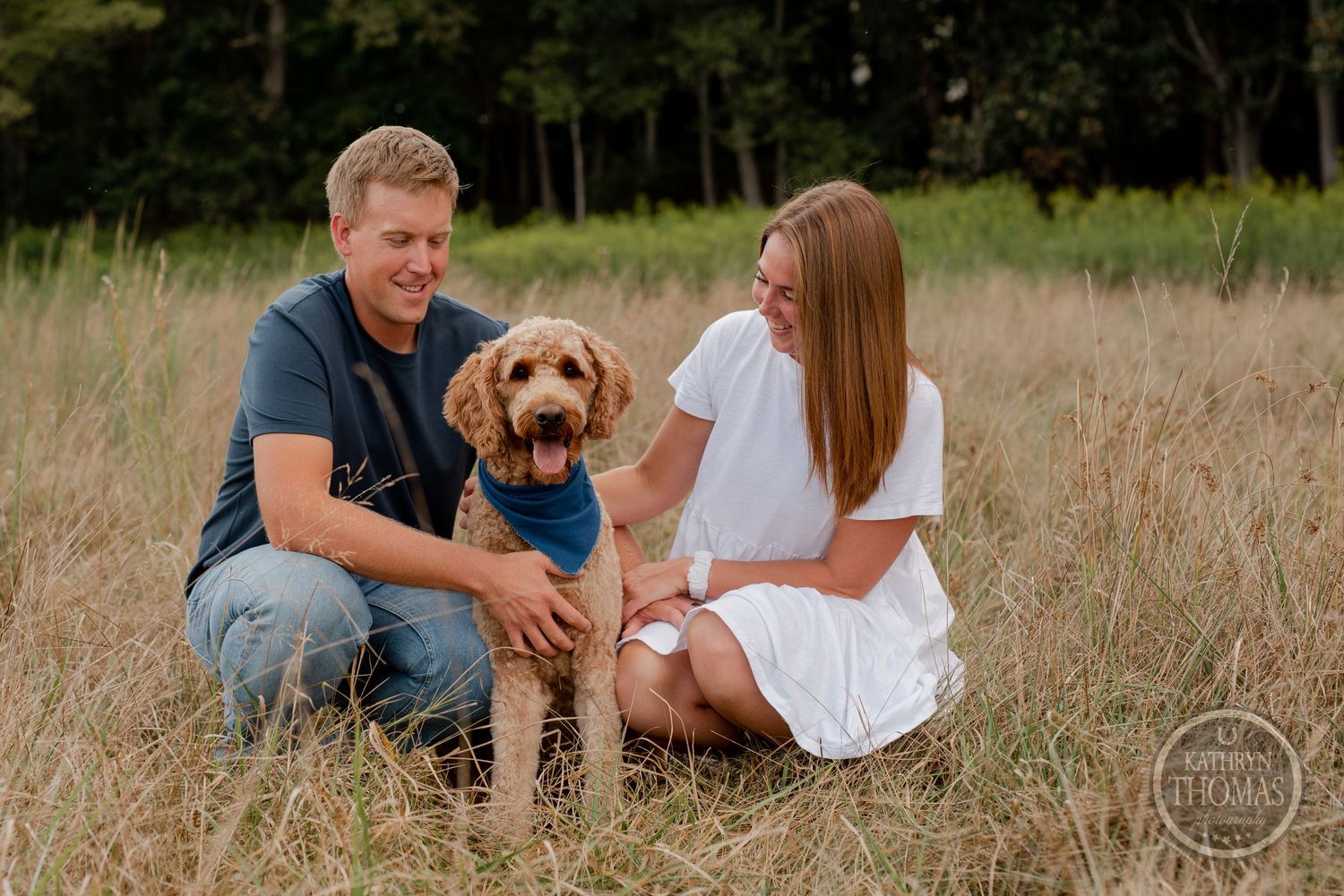 A couple sits in tall grass with their friendly goldendoodle wearing a blue bandana.