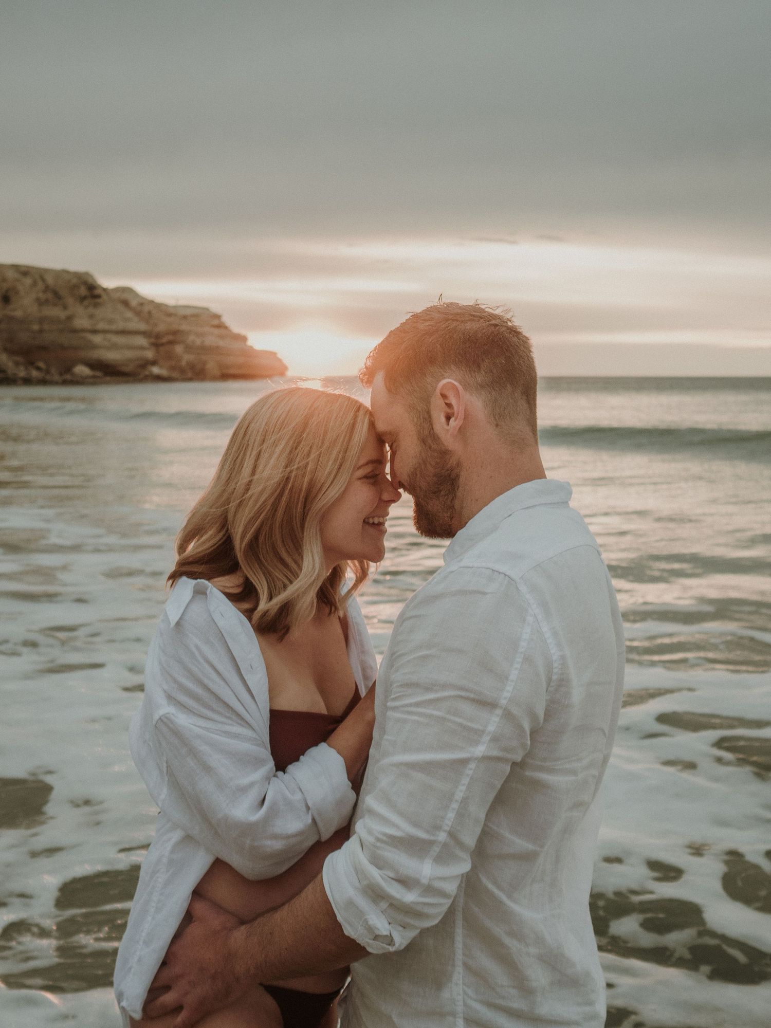 Intimate couple moment on beach with ocean cliffs and sunset backdrop.