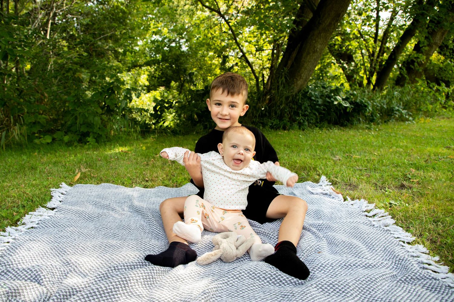 A brother and sister sitting on a blanket with the brother holding his baby sister