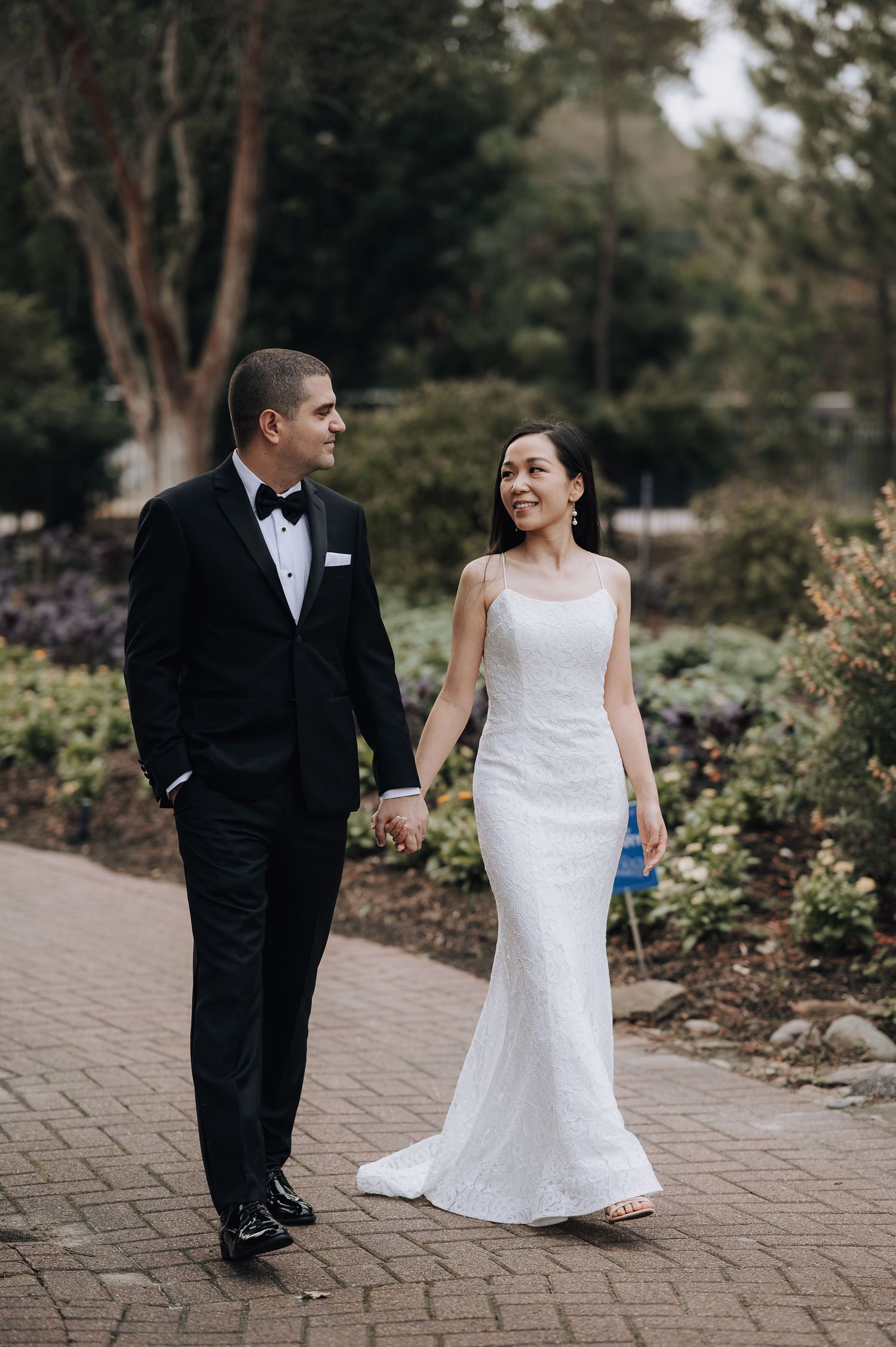 A joyful couple walks hand in hand along a garden path wearing elegant wedding attire.
