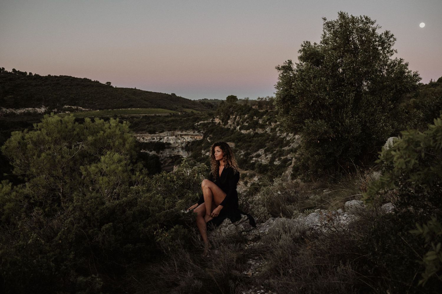 A silhouetted figure sits on a rocky overlook during dusk with mountains in the distance.