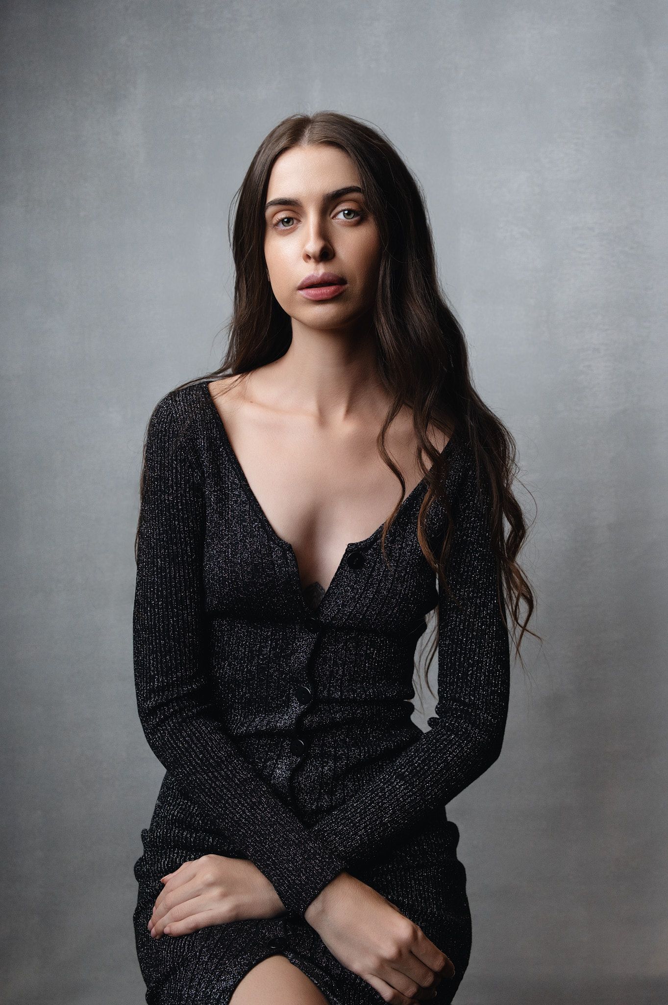 Young woman with long dark hair wearing a black low-cut knit dress, seated against a gray background.
