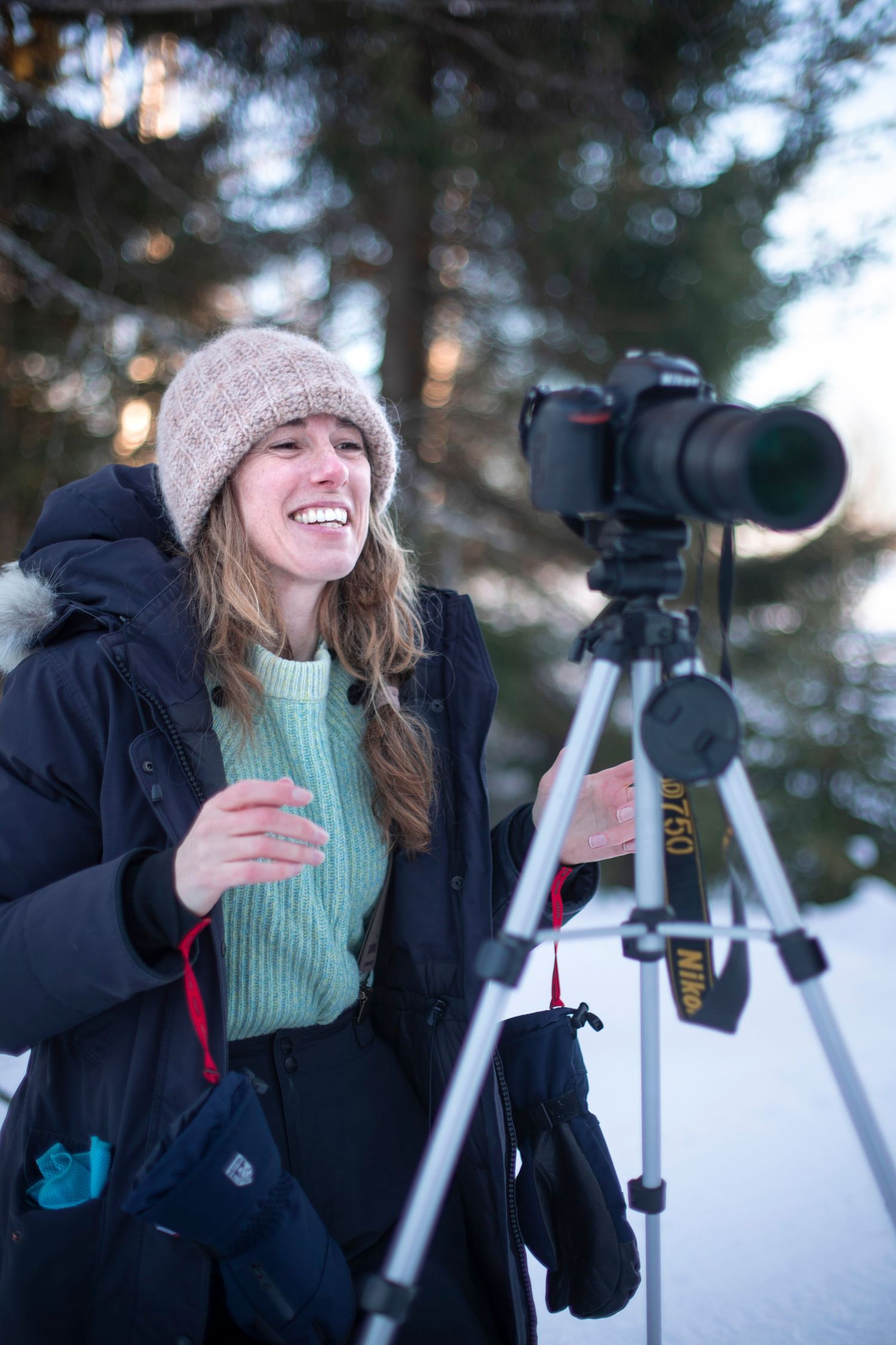 Woman in winter hat laughing beside a Nikon camera on tripod in snowy forest.