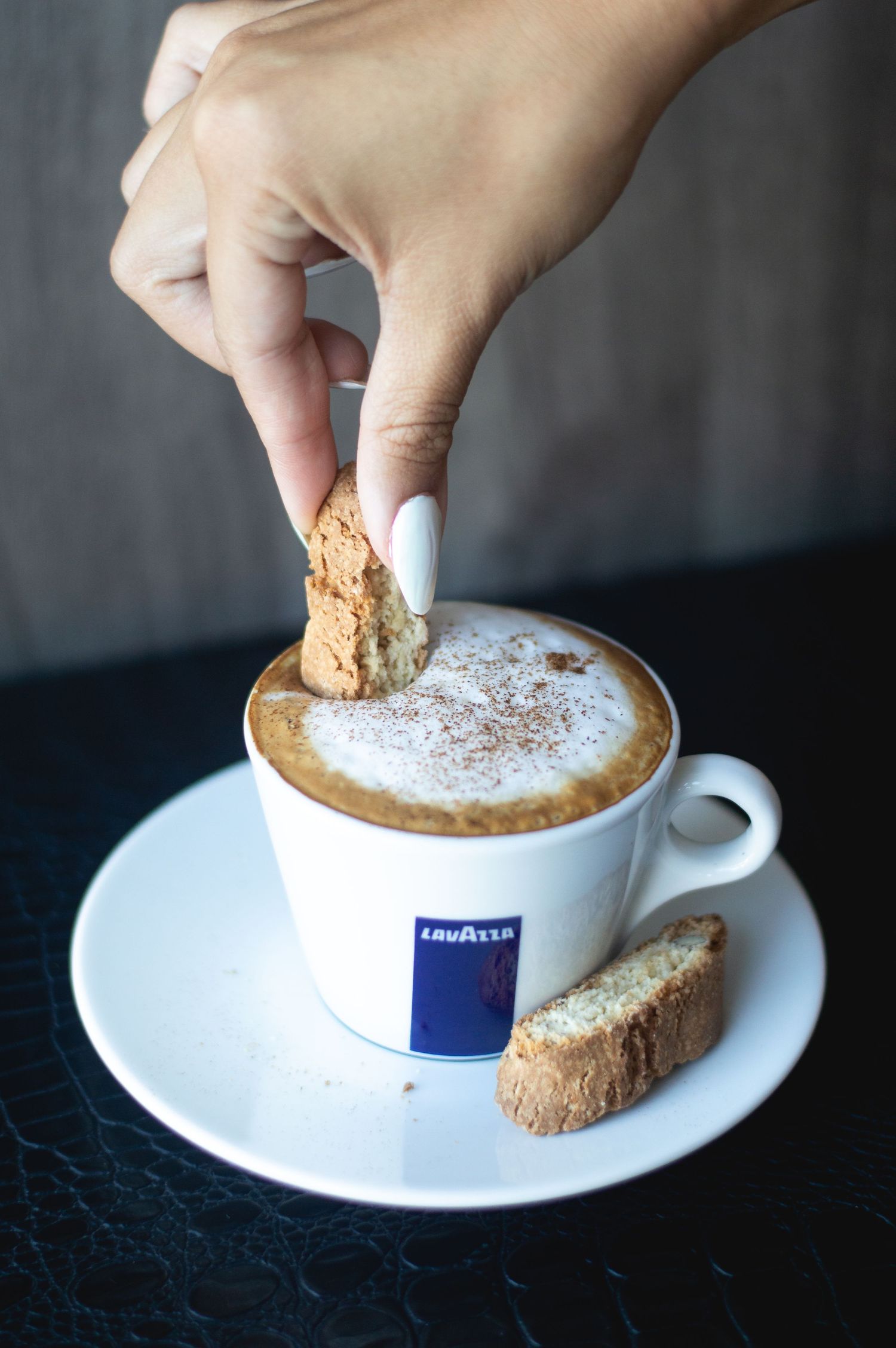 Hand dipping biscotti into a Lavazza cappuccino topped with cocoa powder.