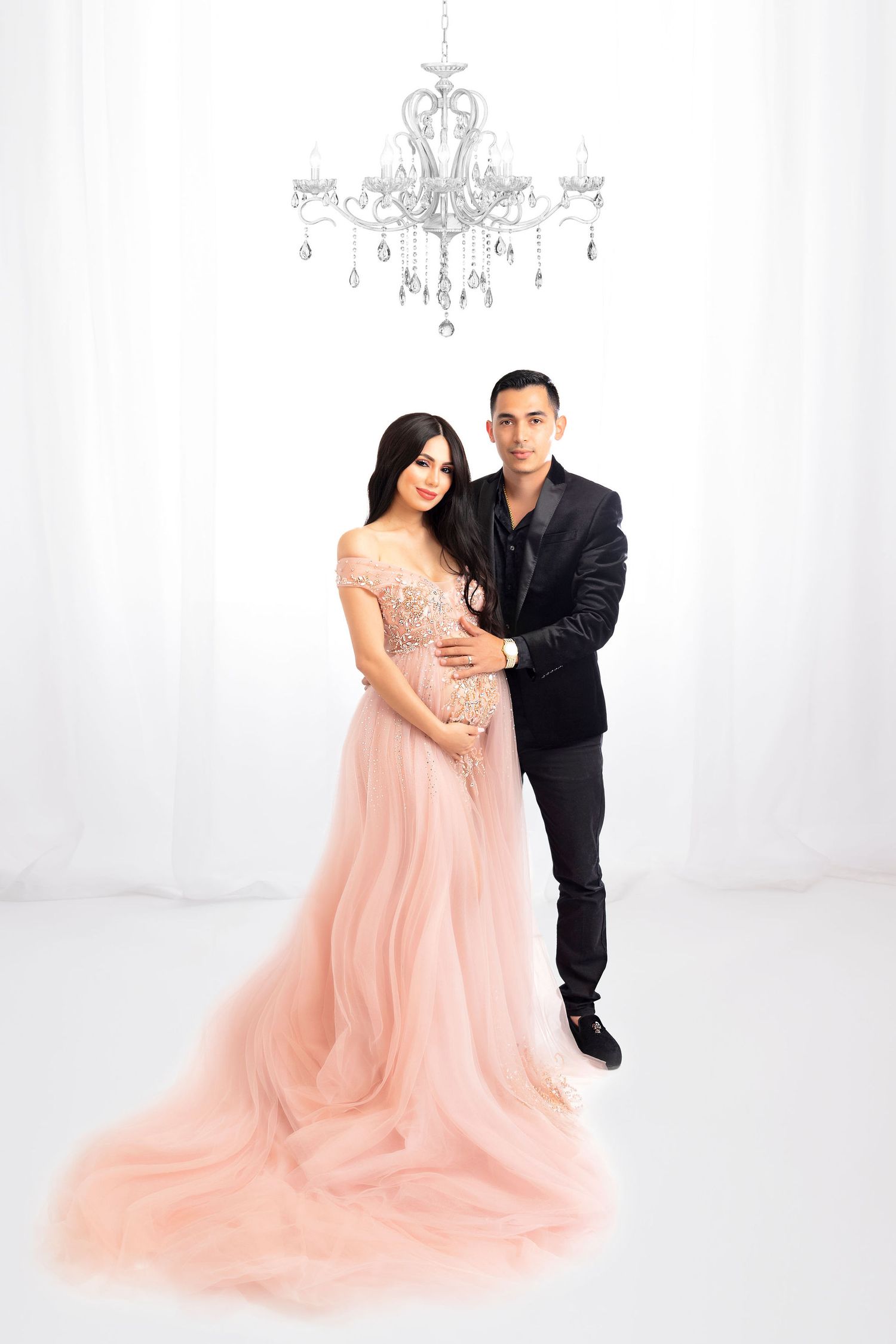 Elegant maternity photoshoot featuring a flowing blush pink tulle gown and crystal chandelier in an all-white studio setting.