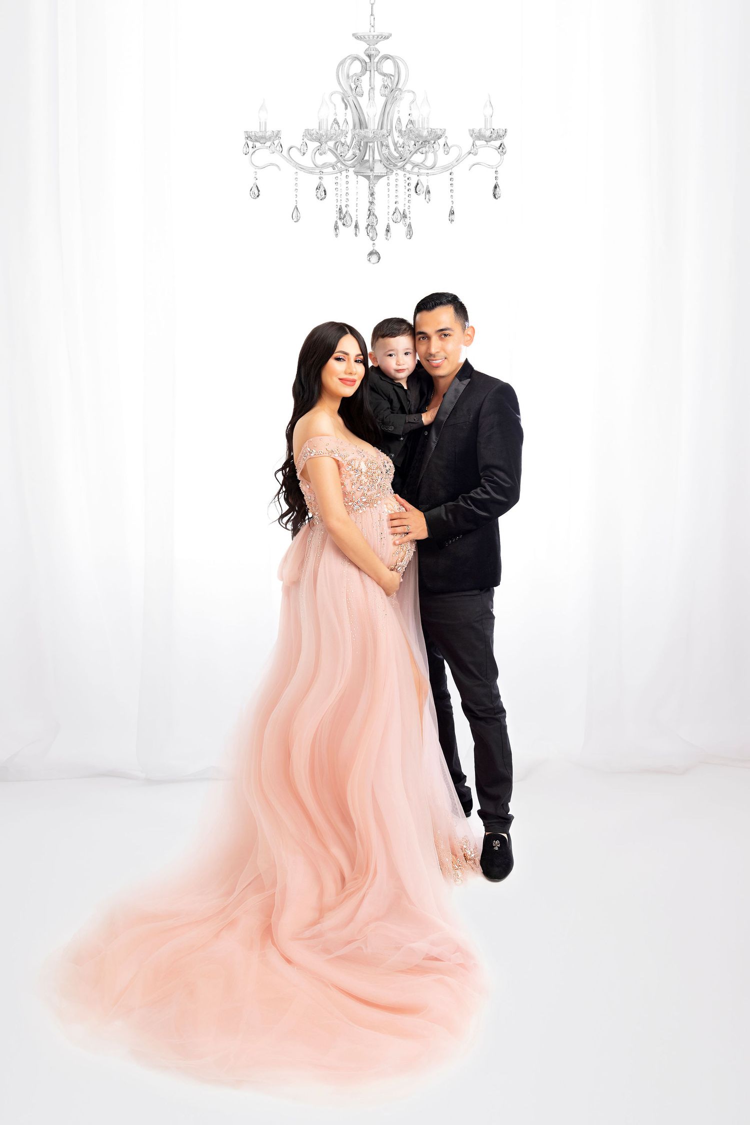 Romantic maternity portrait under a crystal chandelier with a flowing peach-colored gown against white backdrop.