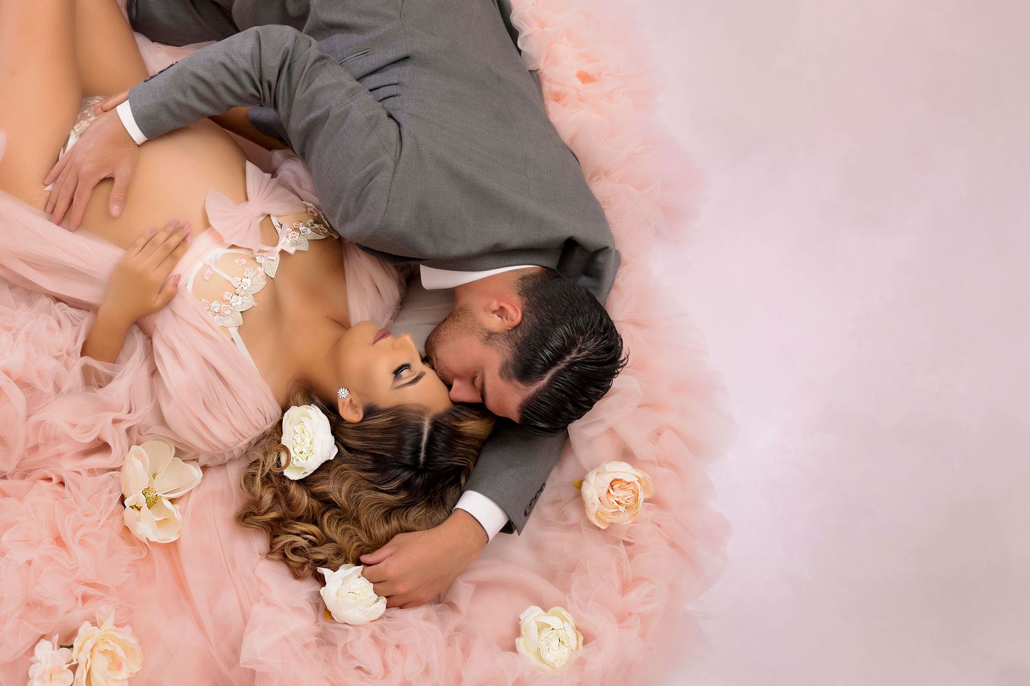 A romantic couple lying on pink tulle fabric with scattered roses during a dreamy photo shoot.