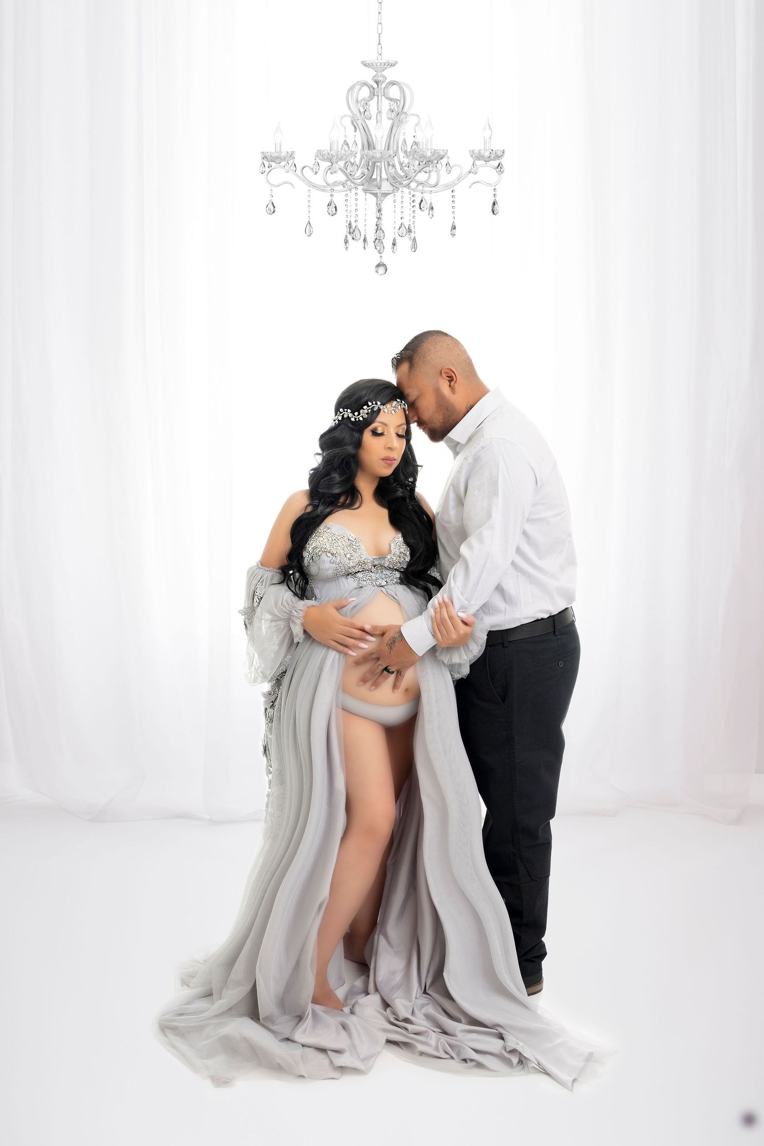 Elegant maternity photoshoot under crystal chandelier with white drapes and formal attire.
