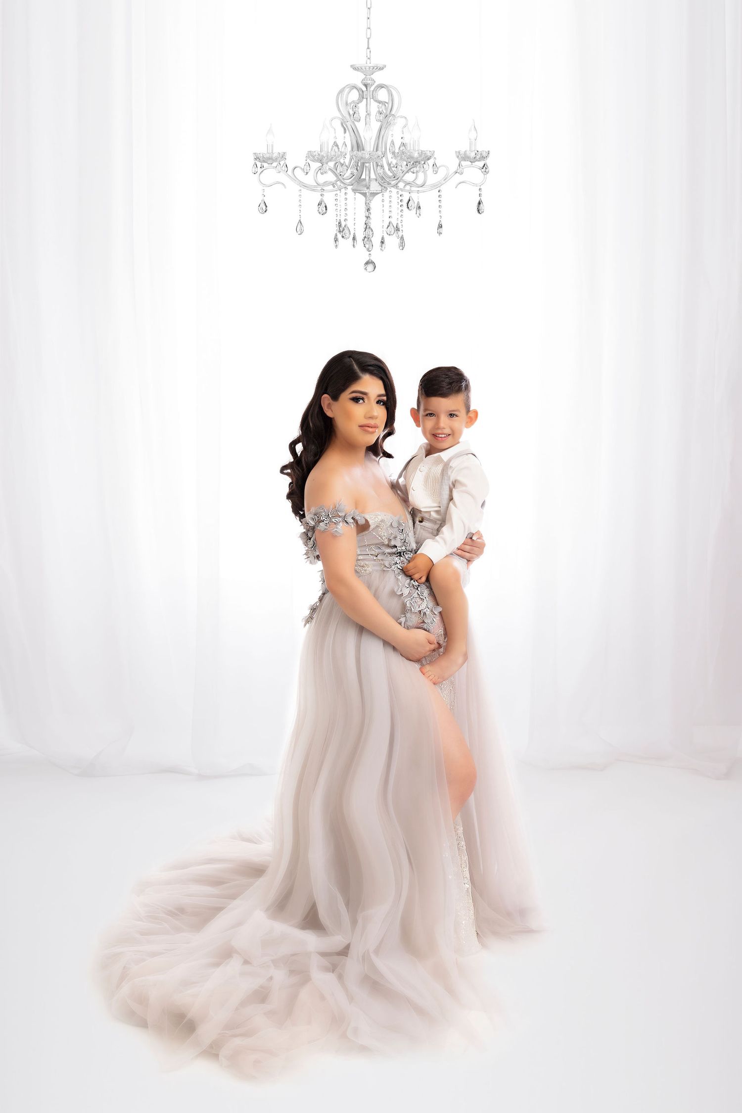 Maternity studio portrait with flowing gray tulle gown and elegant chandelier overhead.
