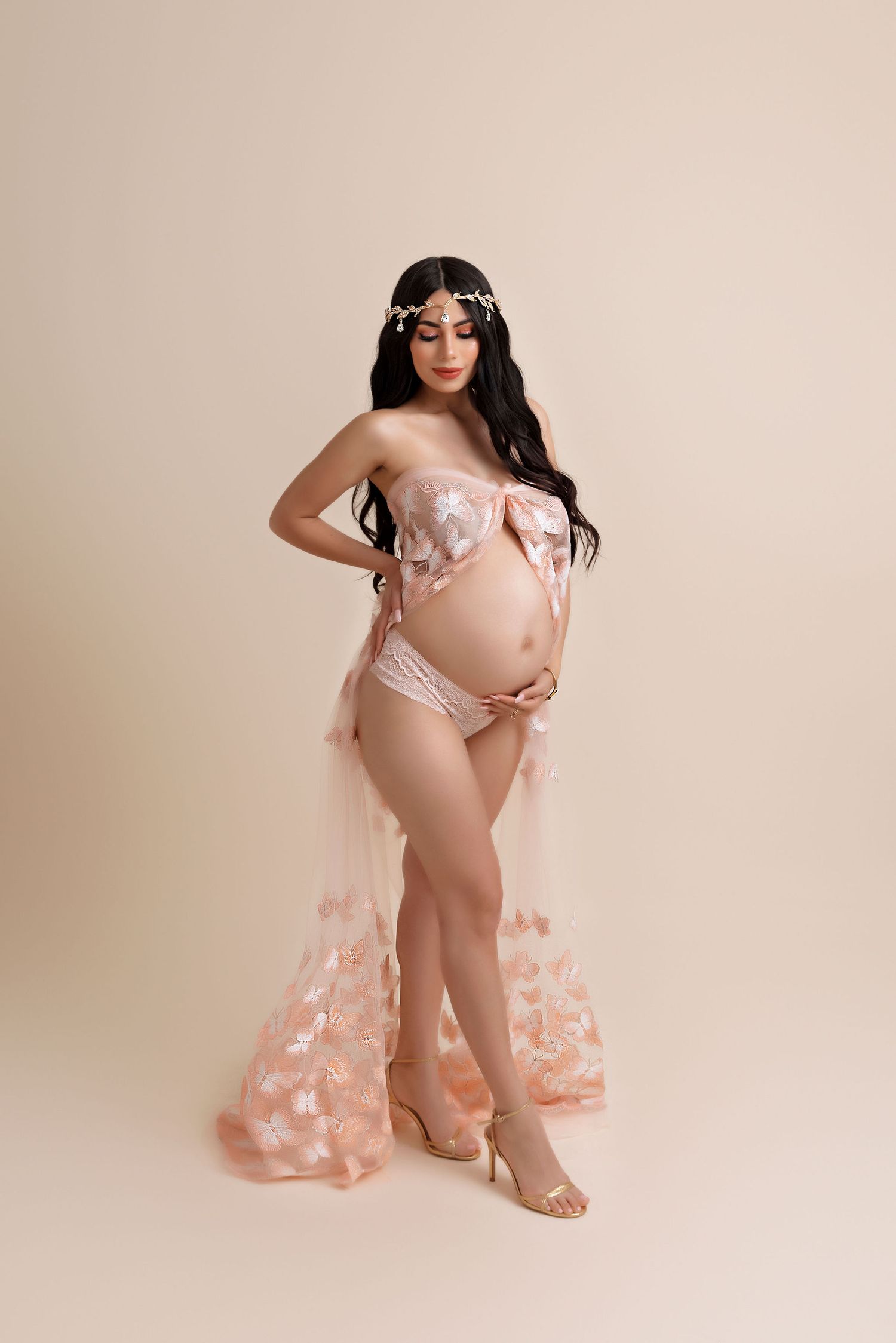 Glamorous maternity boudoir photo with sheer pink floral dress against neutral backdrop.