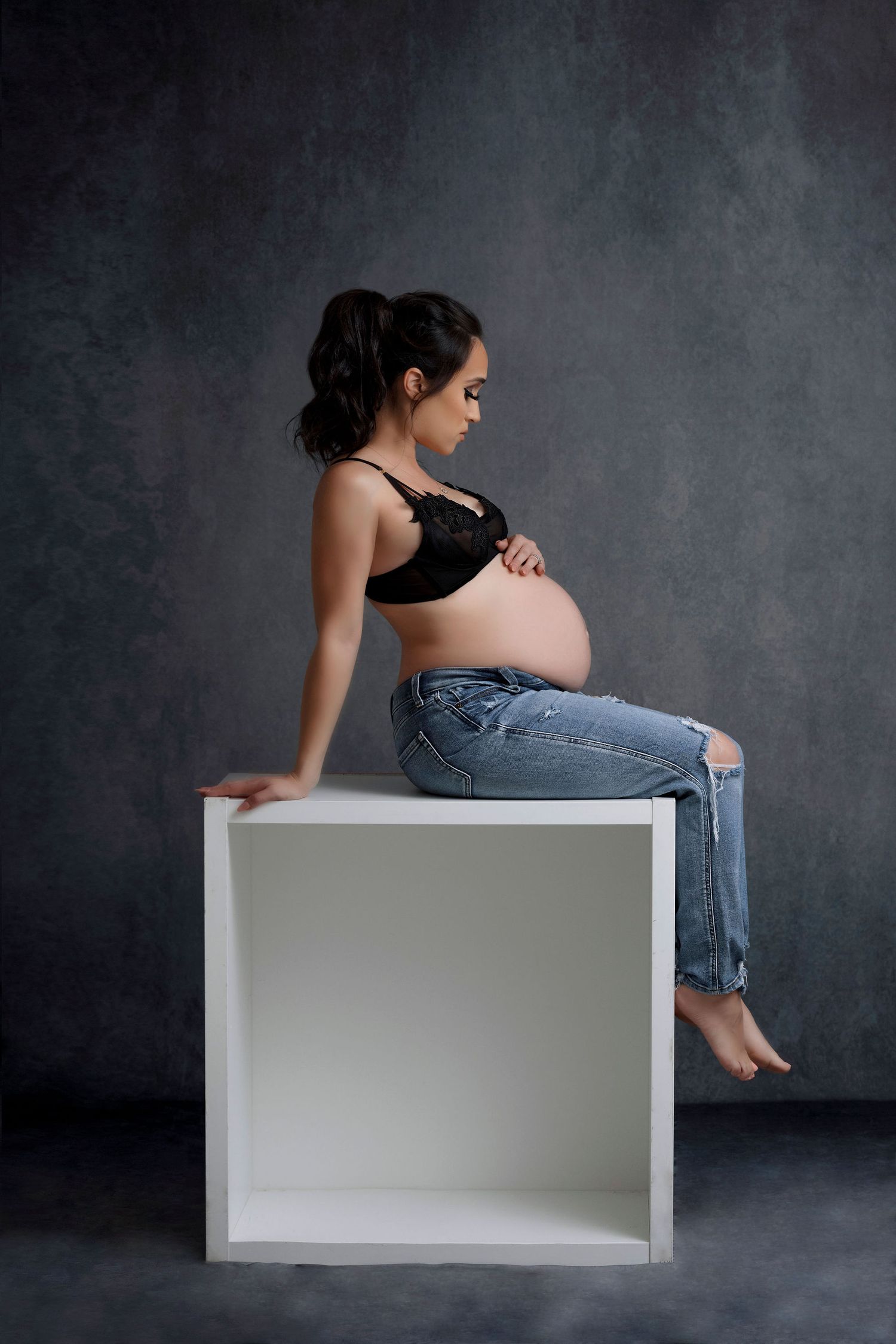 Contemporary maternity portrait with casual denim and black top against dark backdrop.