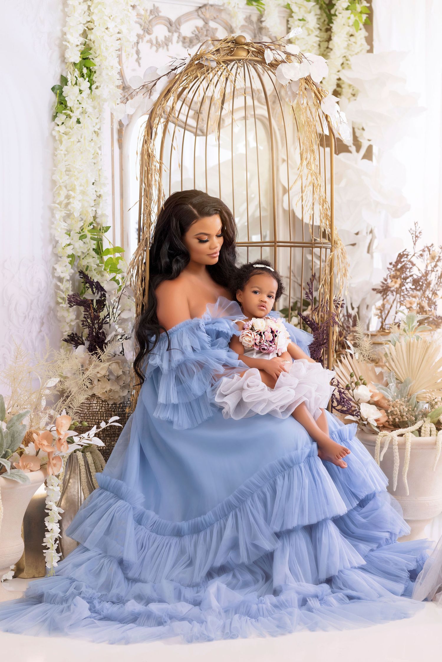 Mother and baby portrait in blue ruffled gown with decorative birdcage and floral backdrop.
