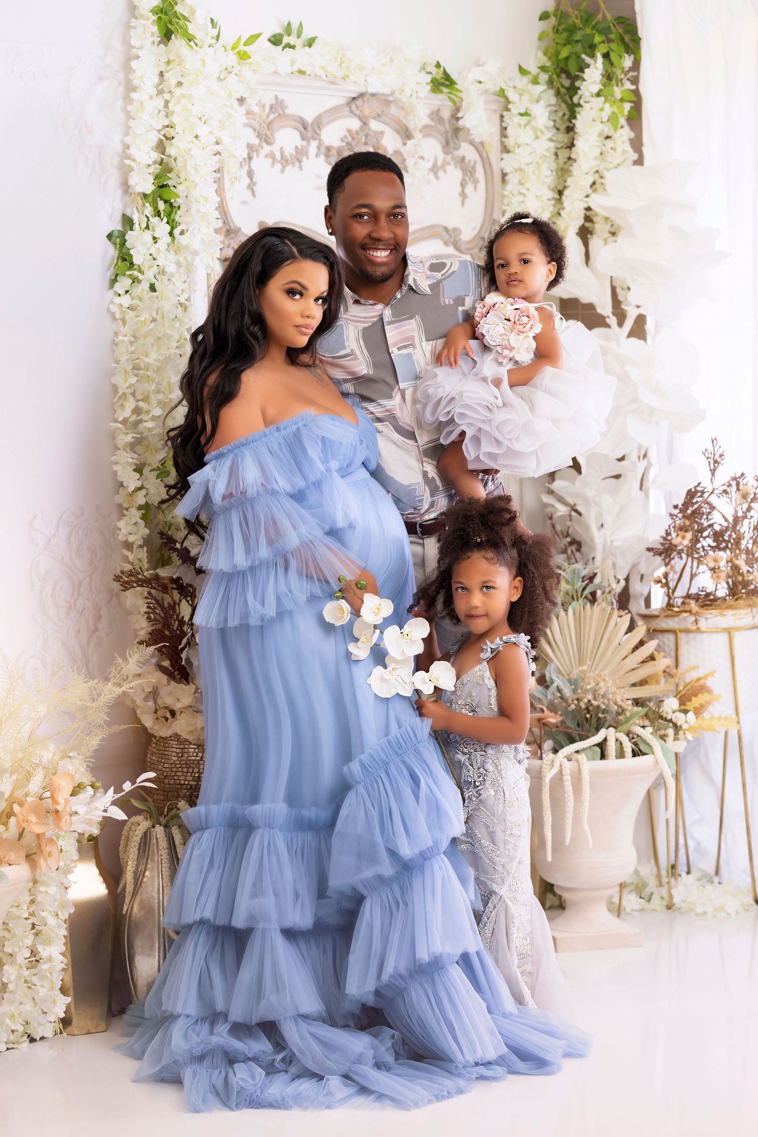 Family portrait with blue ruffled dress against floral backdrop with white decor.