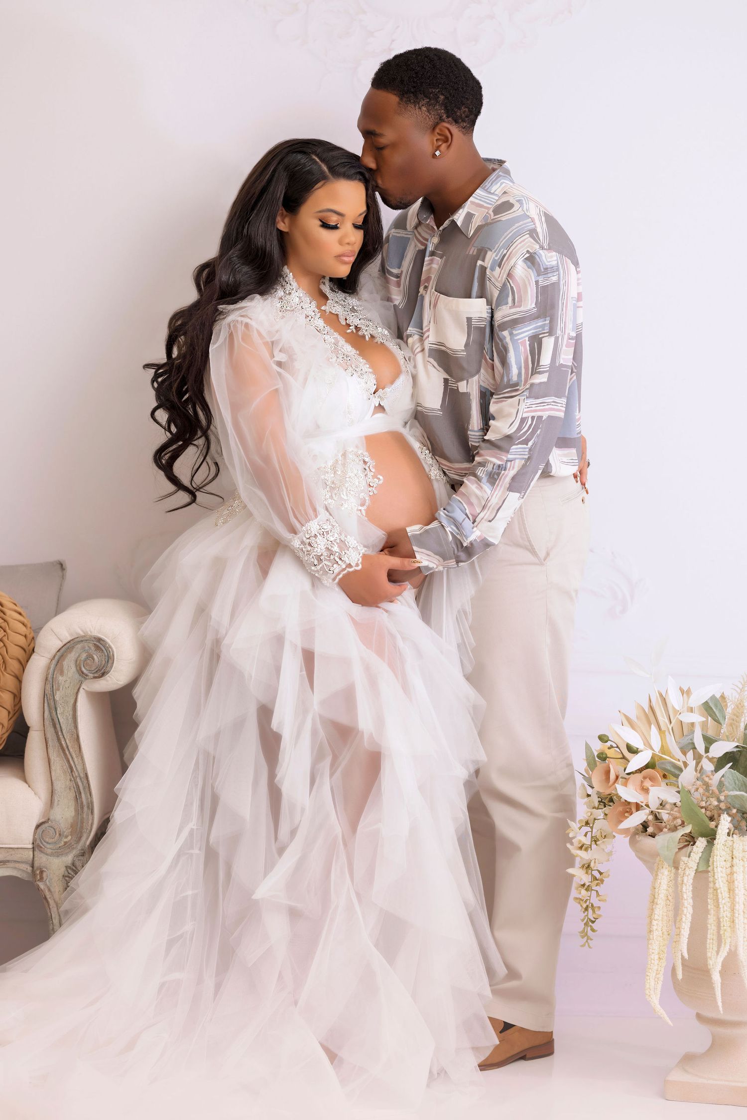 Intimate maternity portrait with couple in white lace dress and patterned shirt against neutral background.