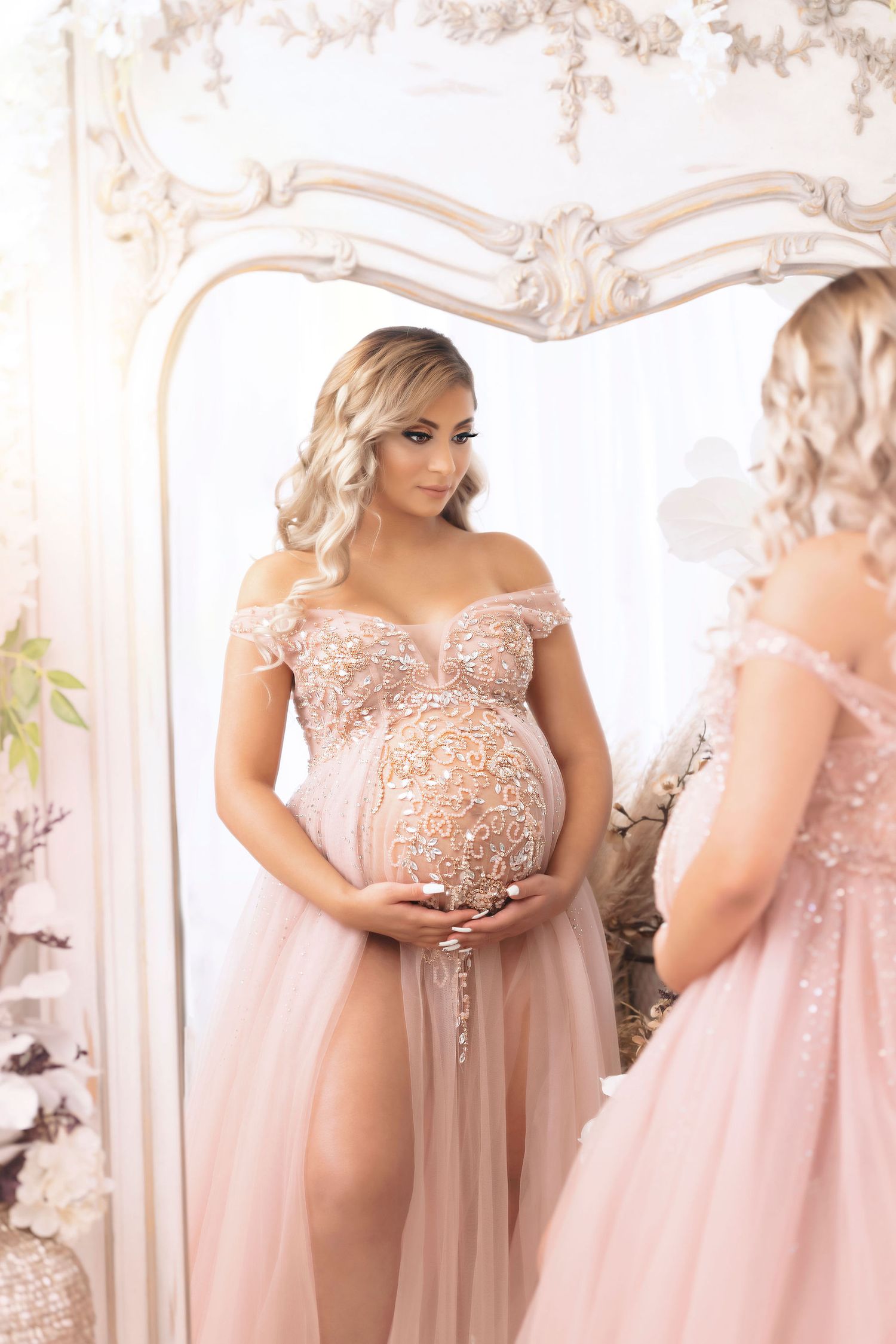 Elegant maternity portrait in blush lace gown reflected in ornate mirror with floral accents.