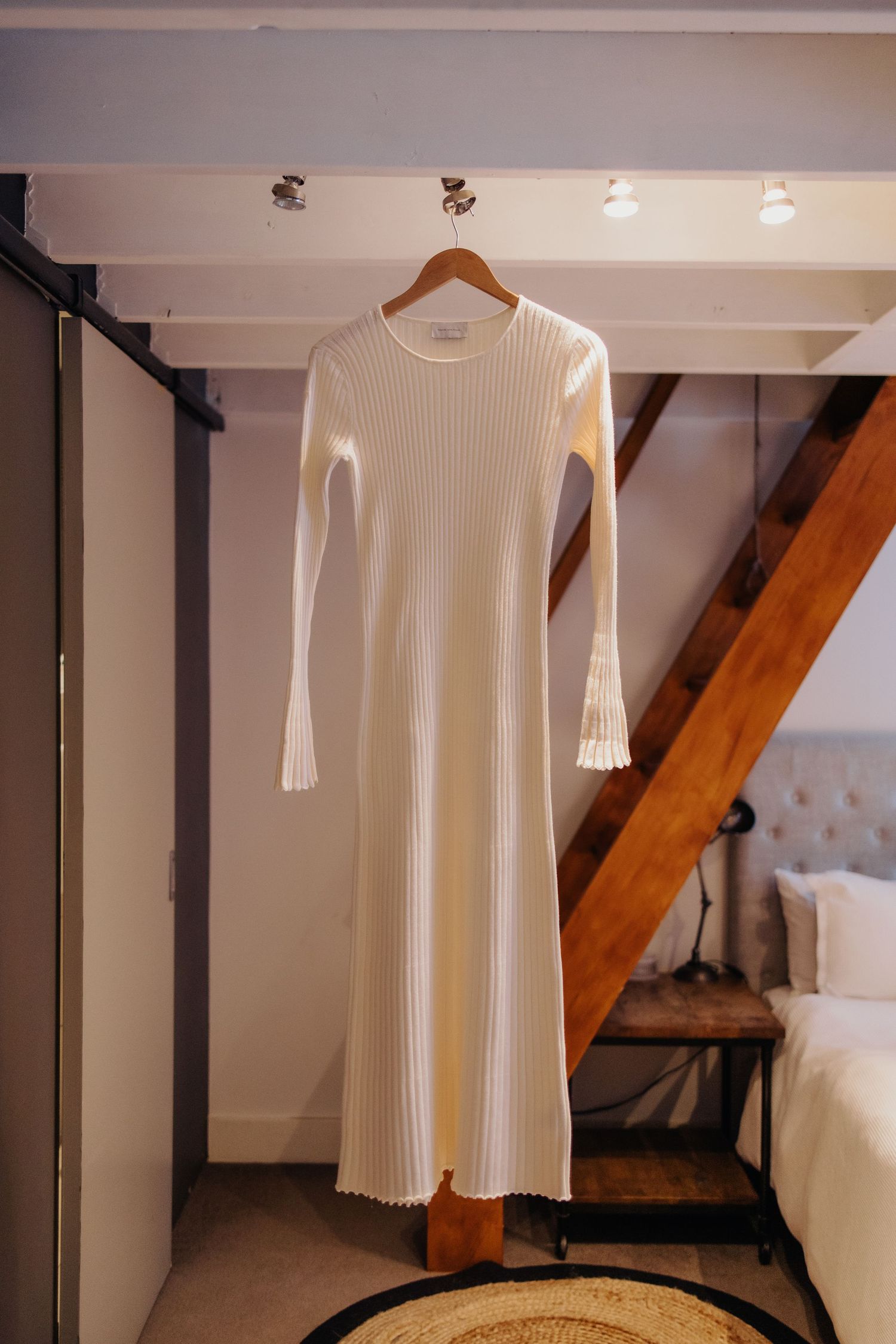 White ribbed wedding dress hanging on wooden hanger in rustic interior setting.