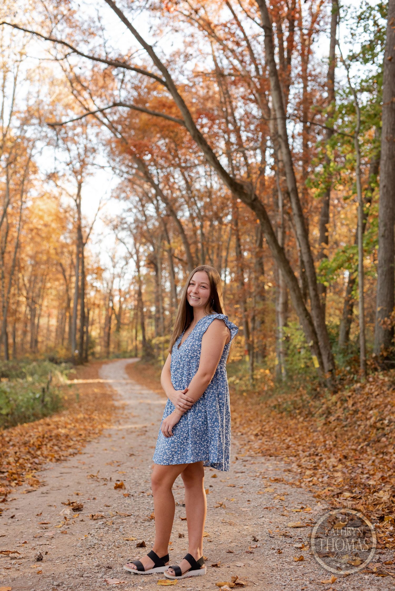 Someone stands on a leaf-covered path wearing a blue dress amid autumn trees with orange leaves.