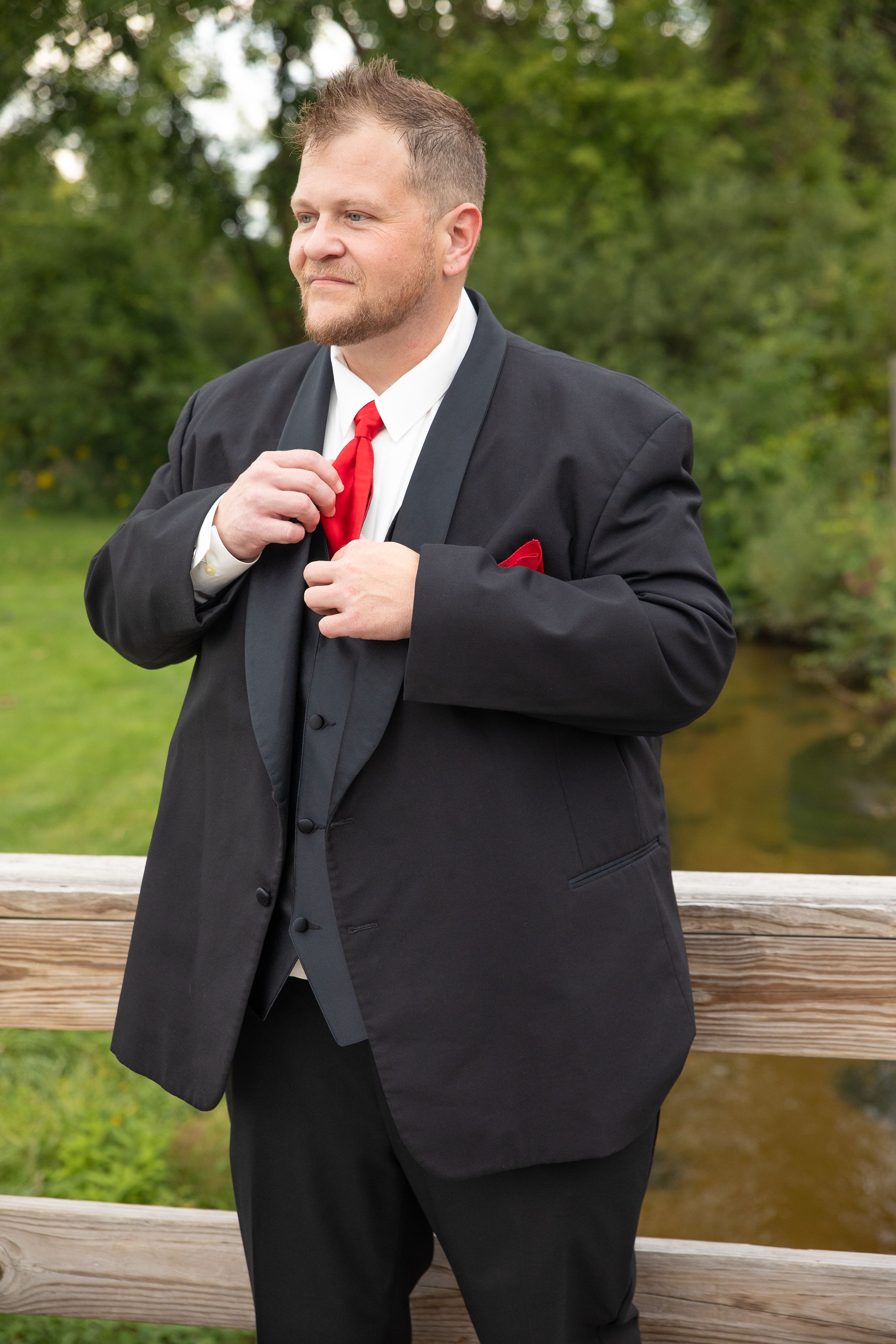 Someone in a black tuxedo with red tie and pocket square adjusts their jacket outdoors.