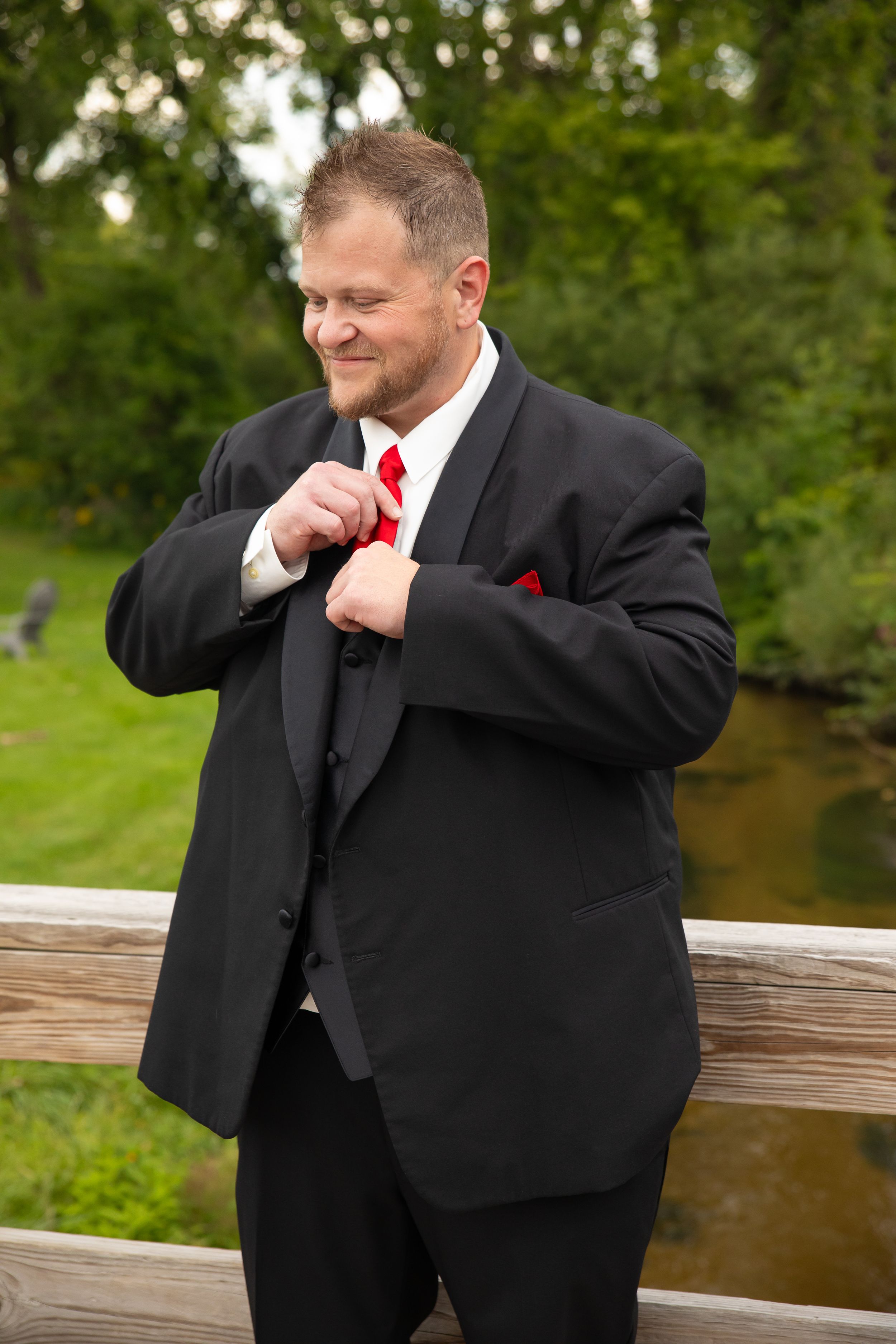 Groom in black suit adjusts his red tie while standing on wooden deck overlooking water.