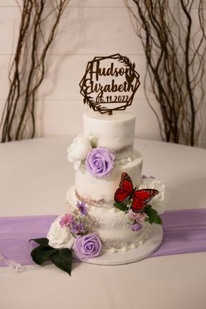 Three-tier white wedding cake decorated with purple roses and red butterfly with custom name topper.